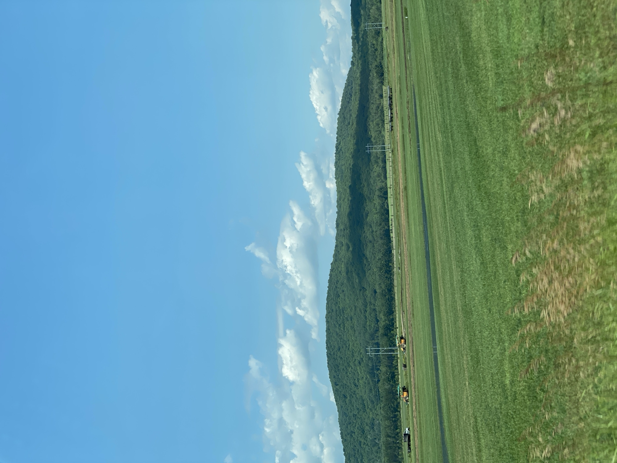 The image depicts a wide, rural landscape under a bright blue sky with scattered white clouds. In the foreground, a vast, vibrant green field stretches horizontally across the frame, with rows of mown grass or crops evident. The midground shows a flatter area of land with some dark, wet-looking patches and what appear to be construction or agricultural vehicles, including yellow excavators and a white truck. Power lines supported by tall poles run across this area. In the background, a densely forested, green hill rises, its contours softened by distance and foliage. The overall scene conveys a sense of open space and agricultural activity. No people are visible. The location is not identifiable as Saint-André-Avellin, Canada, from the image alone.