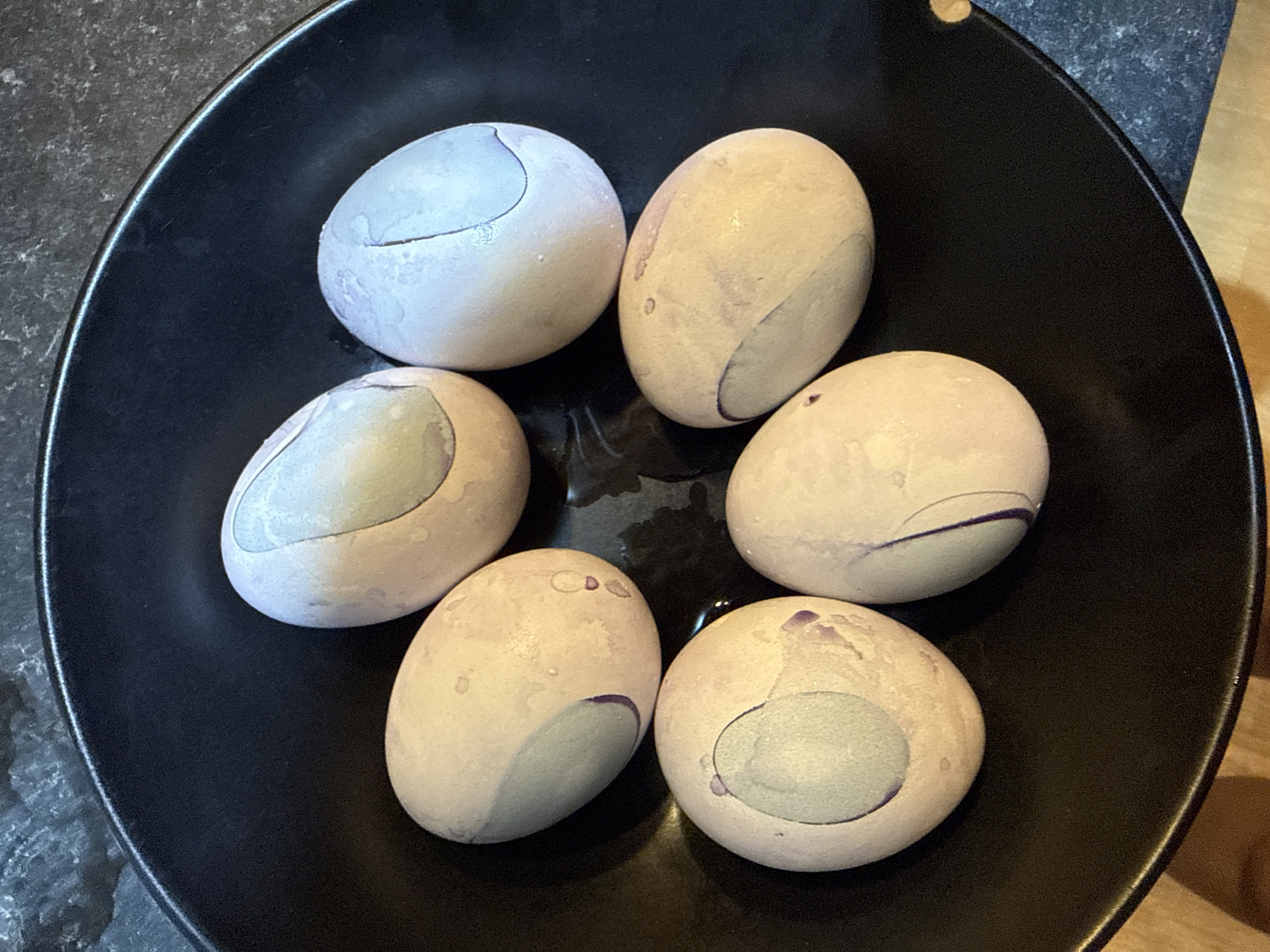 The image shows six cracked and dyed eggs in a black bowl. The eggs have a light-colored base, with portions of the shell having absorbed a gray or blue dye. The bowl is resting on a surface that appears to be part gray stone or granite, and part wood. The location, Great Yarmouth, United Kingdom, is not discernible from the image.