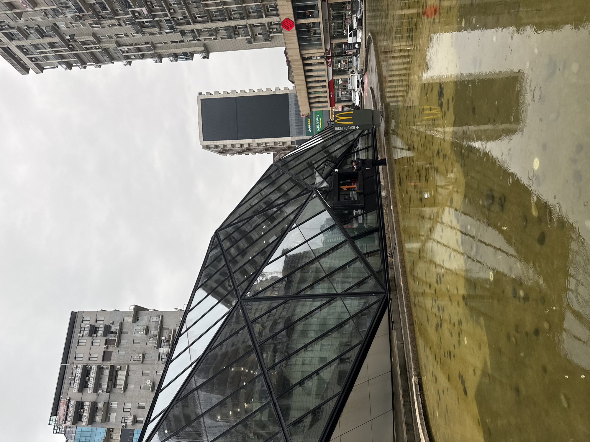 The image captures a street scene in Batumi, Georgia, featuring a modern glass-and-metal structure, potentially an entrance to an underground facility or a distinctive architectural feature. This structure is angled and reflects the surrounding buildings and sky. Adjacent to it is a shallow pool or reflecting pond with a murky yellow-green tint, likely due to the weather conditions and the materials used in the pond.
In the background, several buildings are visible, showcasing a mix of architectural styles. A tall, monolithic building with a large dark screen or panel can be seen, while another building displays a grid-like facade with numerous windows and what appear to be air conditioning units. A McDonald's sign is present, suggesting the presence of a restaurant.
The overall environment suggests a rainy or overcast day, with reflections visible on the wet surfaces and the dull sky. The scene appears to be taken at ground level, capturing the architectural details and the immediate environment.