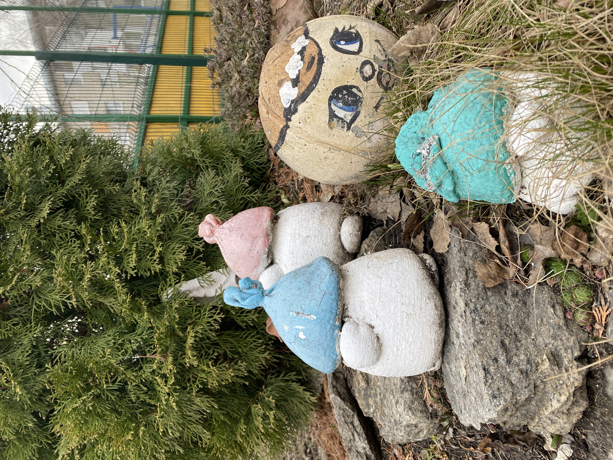 The image depicts a garden or yard setting, likely in Yekaterinburg, Russia given the prompt context. Focus is given to lawn ornaments that are worn or handmade. A stone face painted with cartoonish eyes rests near a grouping of gnome figures. The gnomes have painted caps in various colors (pink, blue, teal) atop white bodies. The area is interspersed with foliage like a coniferous shrub and dried leaves or grass. A fence is also visible in the background.