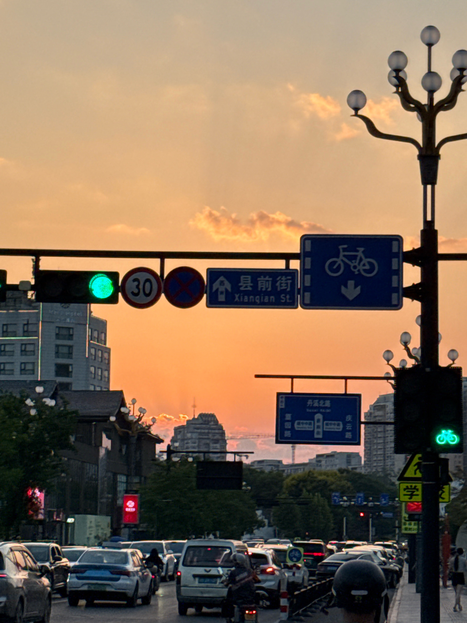 The image depicts a street scene in Yiwu, China, during sunset. The sky is filled with a warm orange glow, with some scattered clouds. Traffic lights are visible, with one showing a green light and another showing a red light. There are several road signs, including a speed limit sign indicating 30, a no parking sign, a street sign for "Xiangqian St.", and a bicycle lane sign with a directional arrow. A decorative street lamp with multiple spherical bulbs stands on the right side of the image. In the foreground, a busy street is filled with cars and motorcycles. Several vehicles are stopped in traffic. People are visible on motorcycles and walking on the sidewalk. Buildings line the street, some appearing modern with multiple stories, while others have more traditional architecture. Trees are also present, adding greenery to the urban landscape.