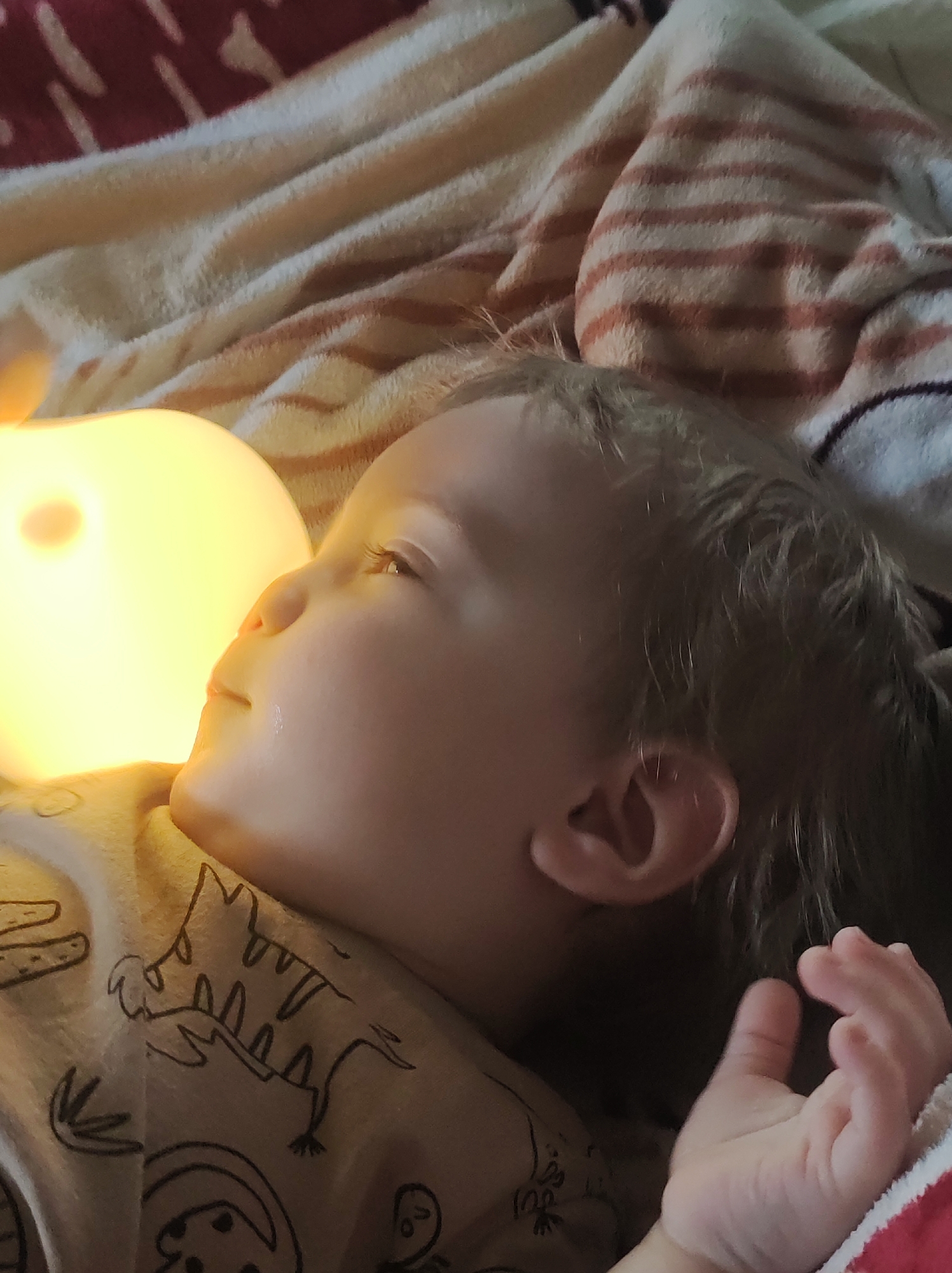 A young child is lying on a striped blanket, illuminated by a nearby glowing orb. The child has light skin and light-colored hair. The child is wearing a shirt with drawings on it. The child's hand is visible. A single tear is visible on the child's face. The location is Unknown, Unknown.