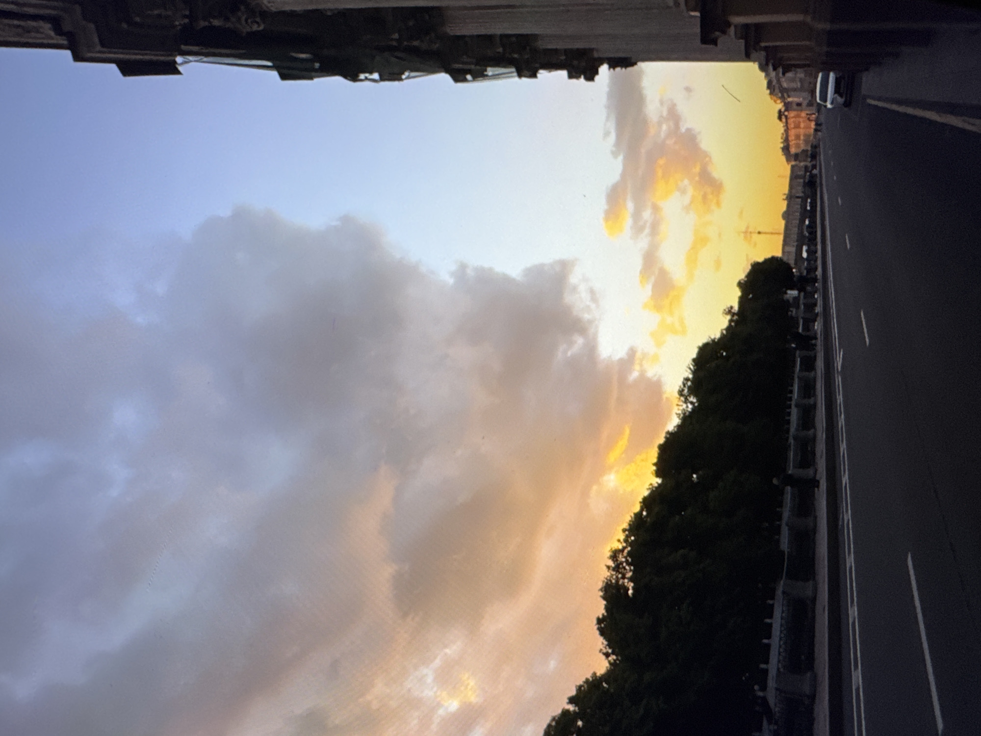 The image depicts a street scene during what appears to be sunset or twilight. The perspective is angled, looking up towards a building facade and across a street. The sky is a prominent feature, with a gradient of soft blues and greys on the left, transitioning to vibrant oranges and yellows on the right, indicating the setting sun. Clouds are scattered throughout the sky, catching the warm light.
On the right side of the image, a dark asphalt road with white lane markings runs parallel to a sidewalk or raised area. A line of trees with dark foliage forms a boundary between the sky and the buildings. Beyond the trees, several buildings are visible, with architectural details suggesting an urban environment. A white car is parked on the side of the road. The overall mood is serene, with the warm hues of the sunset creating a tranquil atmosphere. The time of day is clearly evening, and the weather appears to be clear with scattered clouds. No text is visible in the image.