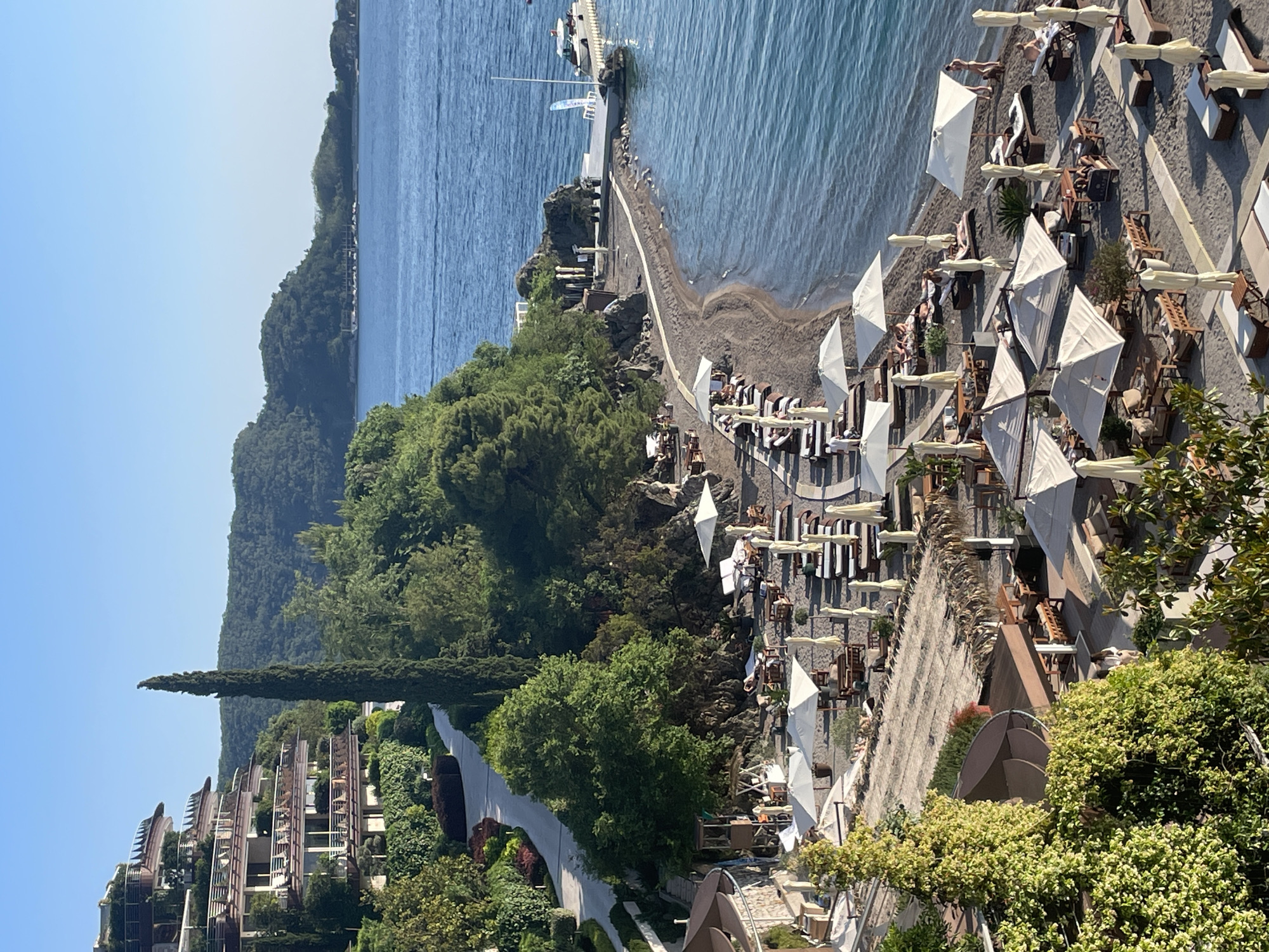 This image shows a beach resort in Budva, Montenegro. The resort features rows of lounge chairs with white umbrellas arranged on a dark sand beach next to clear blue water. People are visible relaxing in the chairs and swimming. A pier stretches into the water with a small boat docked at the end. Lush green trees and landscaping cover the area between the beach and a multi-story building complex with open balconies. A road runs parallel to the resort, and mountains are visible in the background. The sky is clear.