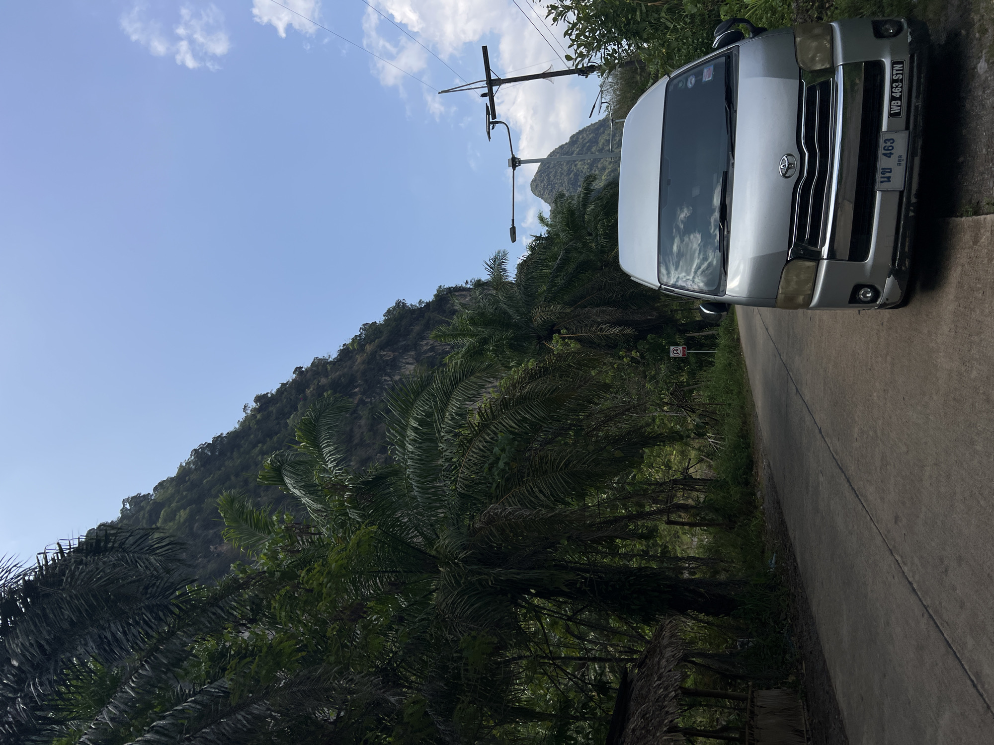 A silver Toyota van is parked on the right side of a concrete road in Ban Nai Wang, Thailand. The van has a license plate that reads "WB 463 STN". Lush green palm trees line the left side of the road, with a mountain rising in the background. The sky is blue with scattered clouds. There is a power pole with wires visible near the top of the frame.