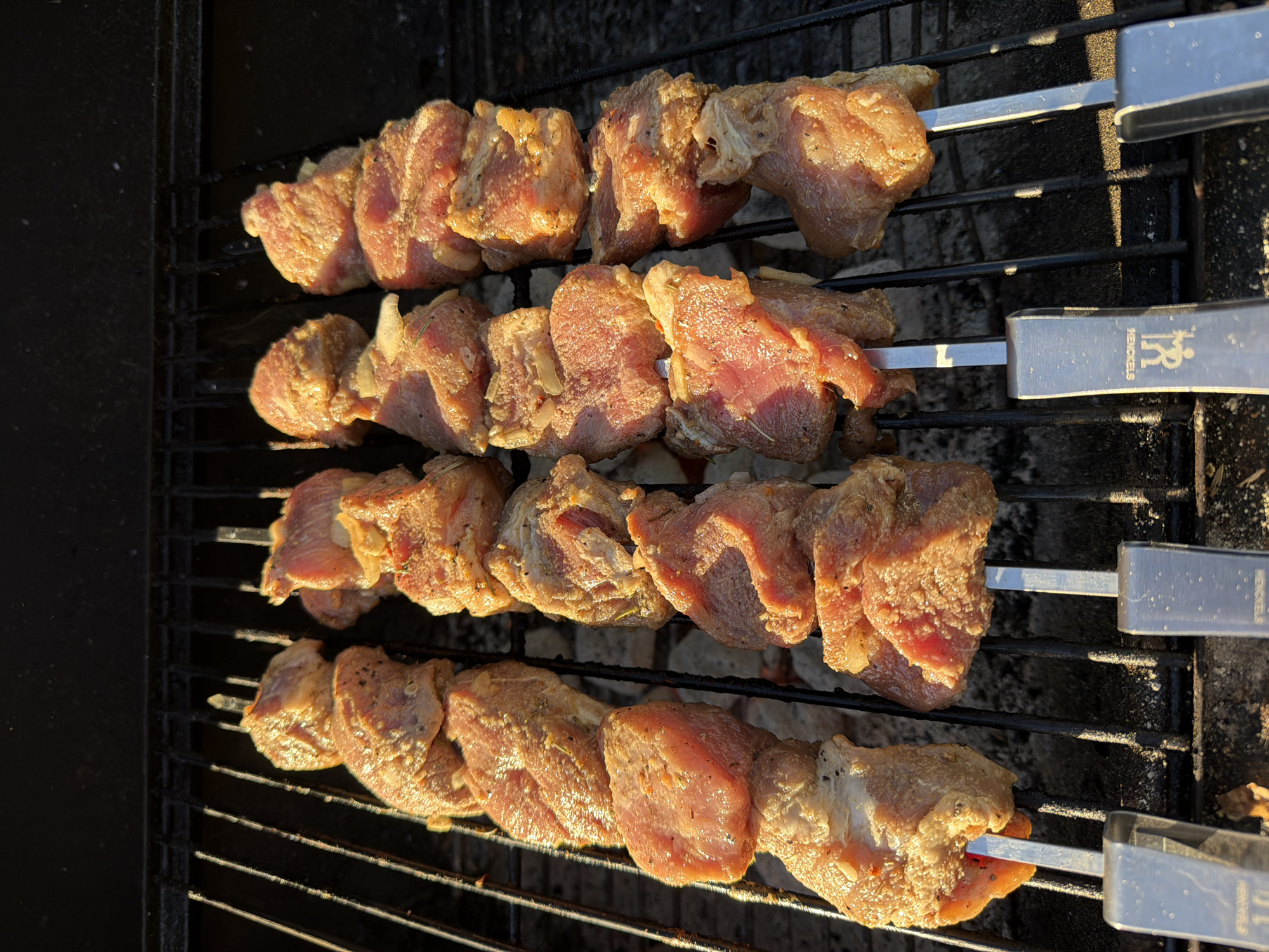 A top-down, eye-level shot shows four metal skewers of raw meat with seasoning and what appears to be some chopped onion on a black metal grill. The meat is cut into uniform cubes. The grill grates are thin and black, and underneath them are greyish-white embers. The scene is illuminated by warm, golden light that casts shadows on the meat and grill. The background is dark and out of focus. The grill is likely outdoors, given the natural light. The image does not contain any information about the city of Sacramento, United States of America.