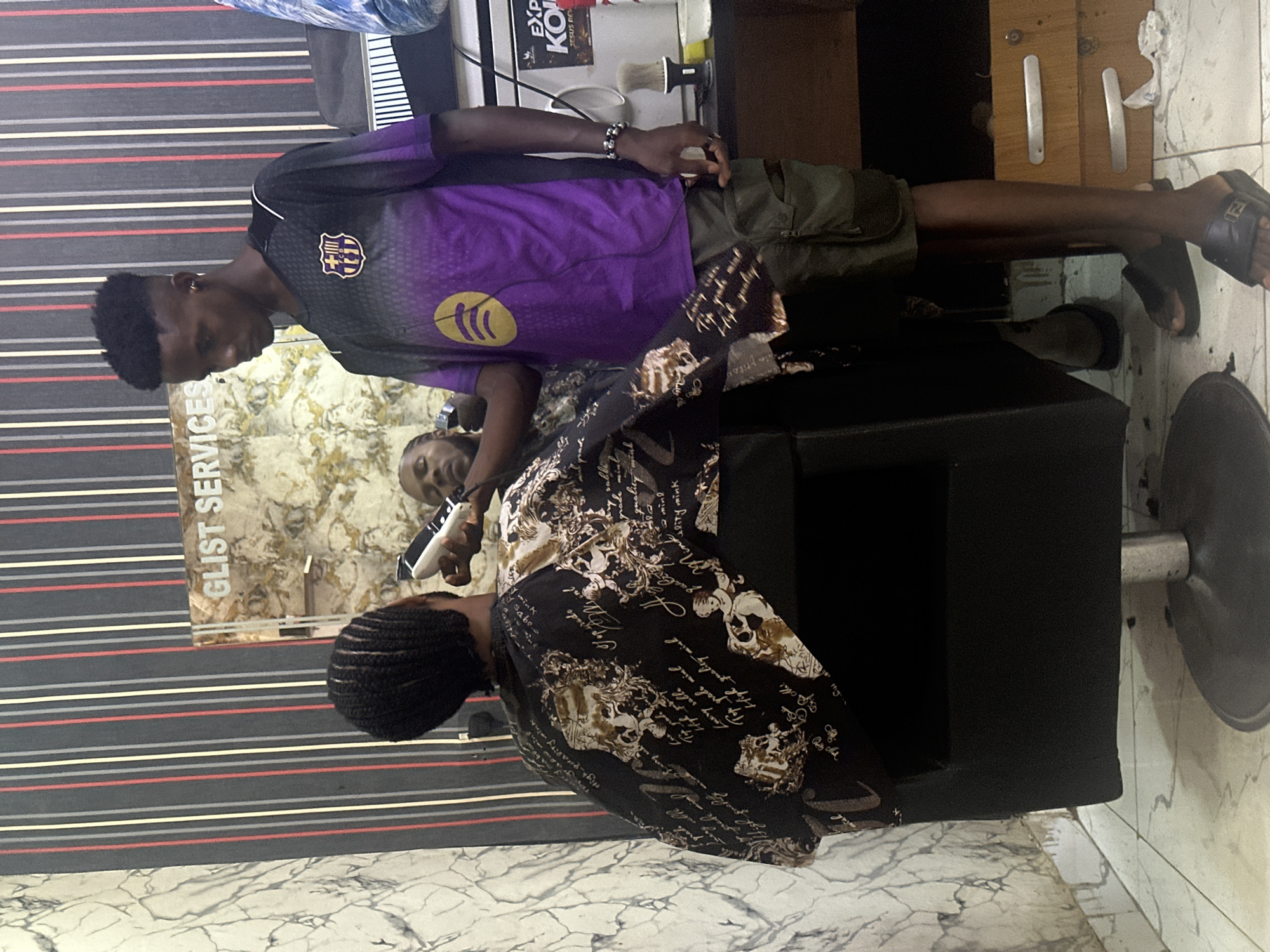 The image depicts a barber giving a haircut to a customer in what appears to be a barbershop in Abuja, Nigeria. The barber, a young man wearing a purple and black FC Barcelona jersey and khaki shorts, is standing and using a pair of clippers to cut the hair of a seated customer. The customer is draped in a black barber's cape with a white and brown print. The barbershop has a striped wallpaper in gray, red, and white, and a marble-like tile floor. An advertisement for "Glist Services" is visible in the background. Other items in the shop include a barber's chair, grooming products, and what appears to be a small refrigerator. The lighting is bright, suggesting an indoor setting.