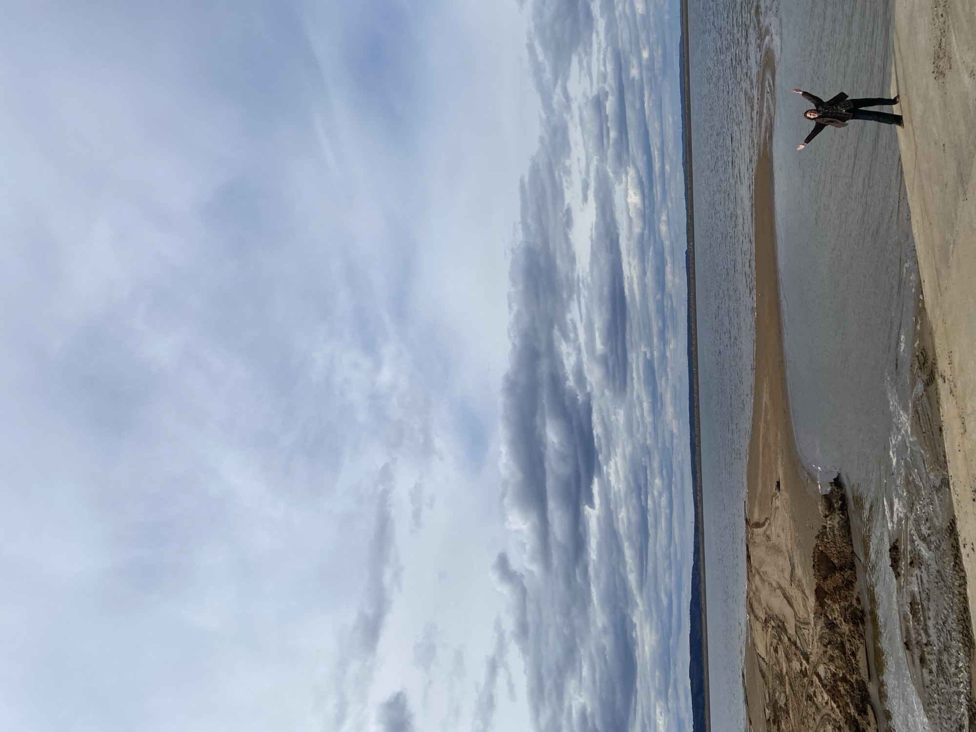 A wide shot captures a person standing on a sandy/muddy bank beside a large body of water under an expansive, cloudy sky. The image is rotated 90 degrees clockwise from its natural orientation.
When viewed correctly, a solitary figure is positioned on the right side of the frame, standing on a slightly elevated bank that slopes down to the water. The person, dressed in dark attire including a jacket and pants, has their arms outstretched, creating a sense of openness or joy against the vast landscape.
The middle ground is dominated by a wide expanse of calm, grey-blue water, resembling a lake or a broad river. A low, distant shoreline or landmass is visible on the far left horizon, suggesting a flat, open environment. The immediate bank where the person stands shows textures of wet sand or mud, leading into the water.
The upper portion of the image features a dramatic sky filled with layers of grey and white clouds. The clouds are arranged in elongated, somewhat parallel formations, creating a textured and dynamic overhead canopy. The light is diffused, indicating an overcast day without direct sunlight. No specific activity beyond standing and posing is depicted. There is no visible text. The scene conveys a sense of quiet grandeur in a natural setting, consistent with a location like Chaikovsky, Russia.