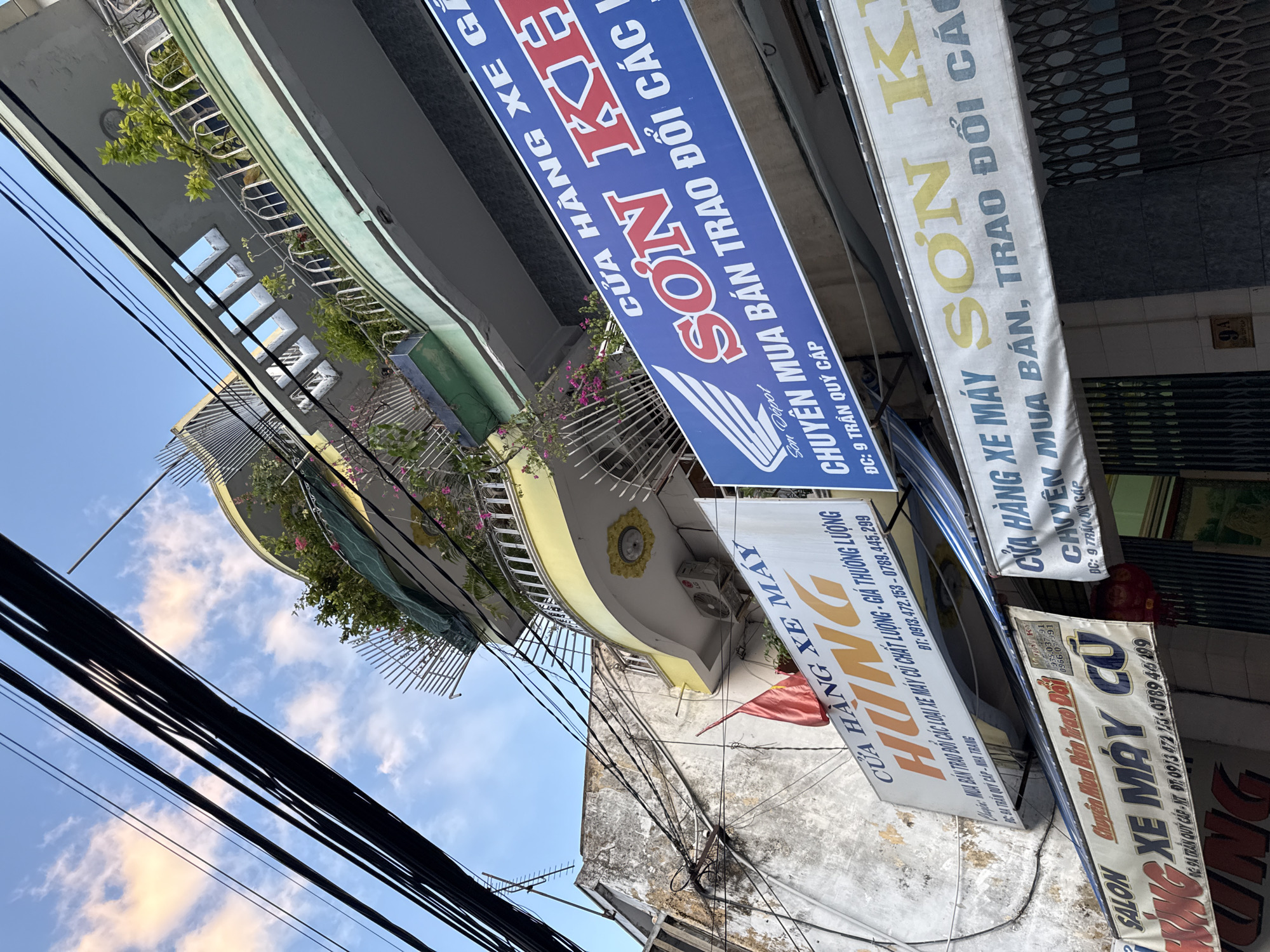 The image captures a street scene in Nha Trang, Vietnam, focusing on the facades of several commercial establishments. The perspective is from below, looking upwards at the buildings and a tangle of overhead electrical wires against a sky with scattered clouds.
One prominent sign, colored blue with white and red lettering, reads "CỬA HÀNG XE GẮN MÁY SƠN KHÔI CHUYÊN MUA BÁN TRAO ĐỔI CÁC LOẠI XE MÁY CŨ." Below this, in smaller text, it indicates "DC: 9 TRẦN QUÝ CÁP." This sign appears to be for a motorcycle dealership specializing in buying, selling, and trading used motorcycles. The Honda logo is also visible on this sign.
Another sign, white with orange and blue lettering, displays "CỬA HÀNG XE MÁY HÙNG." Below the name, it says "MUA BÁN TRAO ĐỔI CÁC LOẠI XE MÁY CŨ, GIÁ THƯƠNG LƯỢNG" followed by a phone number: "ĐT: 0913.472.153 - 0789.445.299." This suggests another motorcycle shop, possibly selling new and used bikes, with a focus on trading and negotiable prices.
A banner below the "HÙNG" sign, with red and blue lettering, repeats similar offerings: "SALON XE MÁY CŨ HÙNG" and "CHUYÊN MUA BÁN TRAO ĐỔI CÁC LOẠI XE MÁY CŨ." It also includes the same phone numbers.
Further to the right, another sign, blue with white text, appears to be "CỬA HÀNG CHUYÊN MUA BÁN XE MÁY CŨ."
The buildings themselves have a weathered appearance, with one particularly old-looking structure to the left showing signs of age and peeling paint. The architecture includes balconies with greenery and railings, some with decorative elements. The overall impression is of a bustling commercial street, typical of Vietnamese cities, with a mix of old and newer buildings and a significant presence of overhead wiring. The street address "9 TRẦN QUÝ CÁP" is a key detail for locating this scene in Nha Trang.
