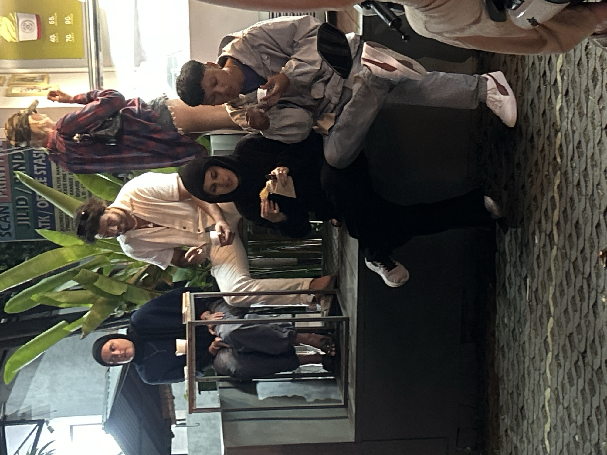 In Kuta Selatan, Indonesia, a group of individuals are gathered in what appears to be an outdoor seating area, possibly a food stall or restaurant, illuminated by artificial lighting. One person, wearing a dark headscarf and dark clothing, sits on a bench with a small white cup and a packaged food item. To their right, another person in a black headscarf and dark clothing holds a similar food item.
Further to the right, a person in a light-colored, open-collared shirt and white pants is seated, holding a white cup. Next to them, a person in a dark, plaid long-sleeved shirt and denim shorts, wearing a camouflage baseball cap, stands and appears to be interacting with something in their hand. Another individual, wearing a light gray hooded jacket and jeans, is seated and holding a small white cup, eating from it. In the foreground, a person's lower legs and feet are visible, wearing white sneakers with red accents. The ground is paved with a textured, interlocking material. Lush green plants are visible in the background, and a sign with text is partially obscured.
