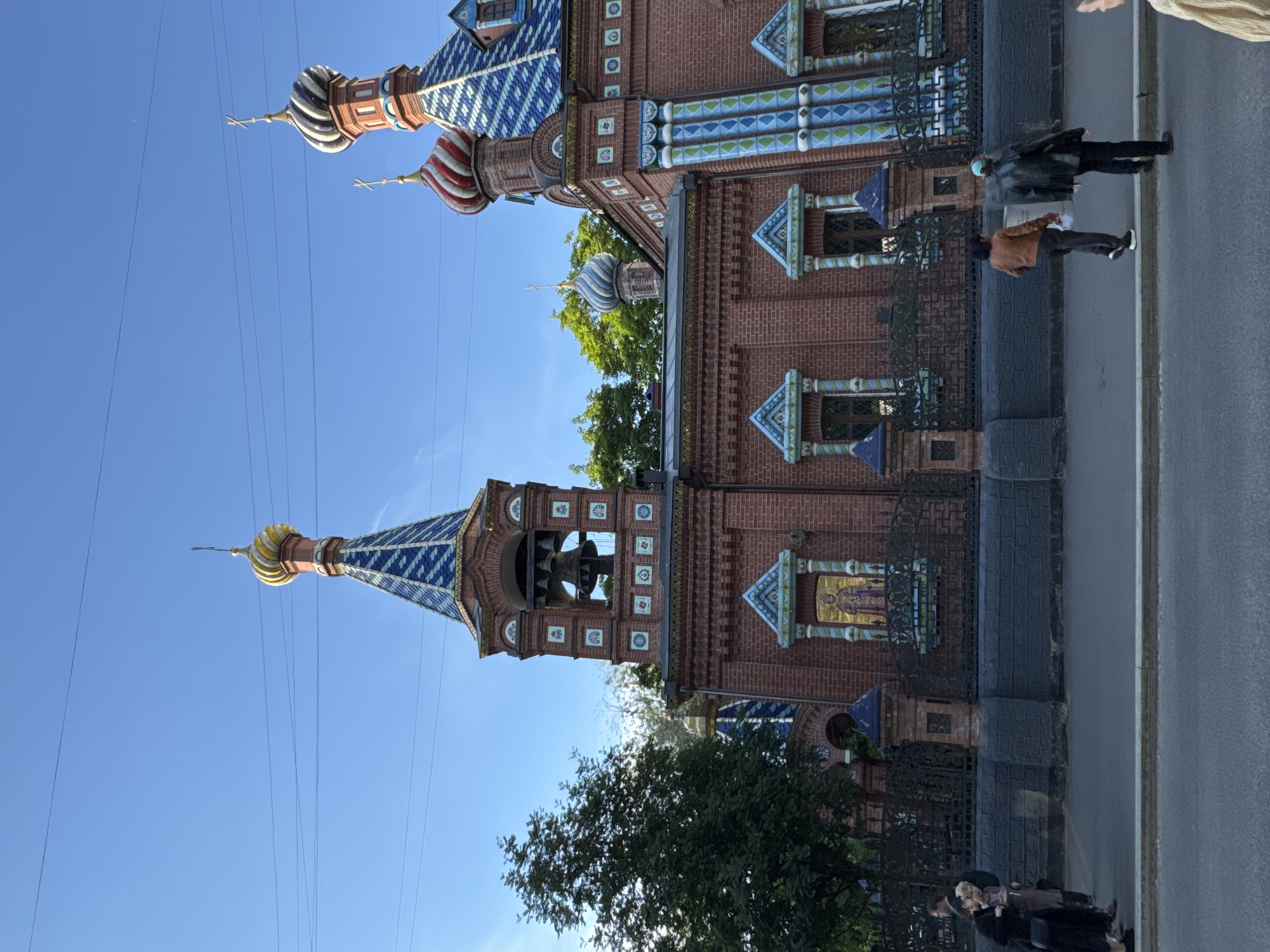 The image depicts an ornate Russian Orthodox church or cathedral under a bright, clear blue sky. The building is primarily constructed of red brick, adorned with richly colored glazed tiles in patterns of blue, white, green, and gold, decorating its rooflines, cornices, and window surrounds. Several onion domes are visible, including distinctive red and white striped, blue and white striped, and gilded variations. A bell tower, featuring visible bells, stands prominently on the left side of the structure, topped with a gilded onion dome. Mosaic icons, depicting religious figures, are integrated into the facade of the building.
A black decorative metal fence runs along the base of the church, separating it from a paved sidewalk and street. Green trees are present behind and beside the building. Overhead utility wires crisscross the expanse of the blue sky.
Several people are seen on the sidewalk. One individual in a brown jacket and dark pants walks away from the viewer, while another person in a dark jacket and pants, possibly wearing a blue hat, walks towards the viewer. Additional people are partially visible further along the sidewalk.
The scene takes place during daytime, characterized by bright sunshine and the unclouded sky. The distinctive architectural style, including the onion domes and vibrant tiling, serves as a strong location cue for Moscow, Russia. No discernible text is visible within the image.