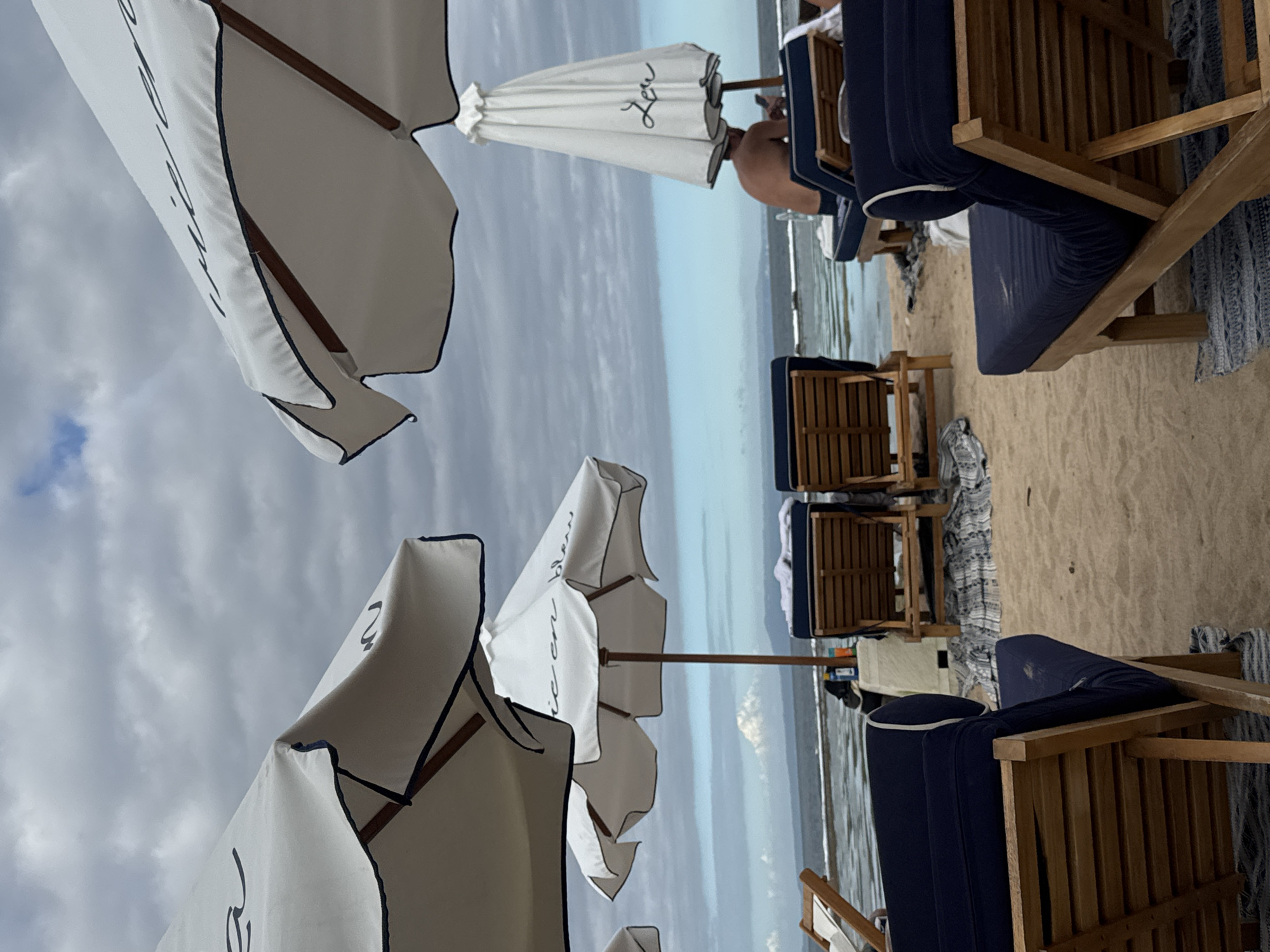 The image depicts a beach scene in Kuta Selatan, Indonesia, featuring beach umbrellas and lounge chairs. The foreground shows white beach umbrellas with dark blue trim, angled towards the camera. Behind them, lounge chairs with dark blue cushions are arranged on the sand. One person is visible reclining on a chair, partially obscured by an umbrella. The background shows the ocean and a cloudy sky.