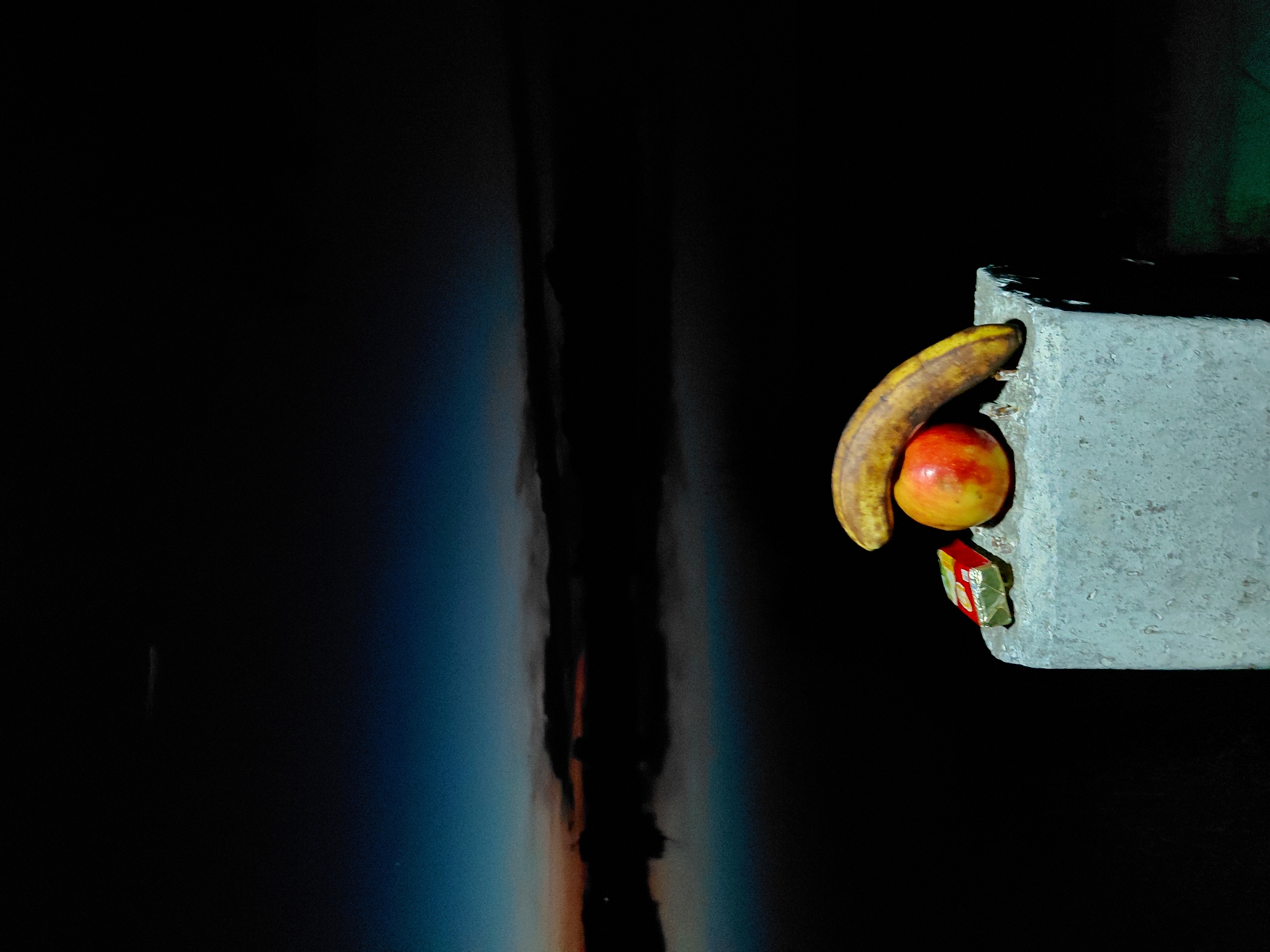 A still life composition is presented on a rough, concrete surface against a dark, out-of-focus background. The concrete block, positioned in the lower right quadrant of the frame, has an uneven texture with visible imperfections. Resting on this surface are three items: a red apple with yellow and orange streaks, a slightly curved, yellow banana, and a small, rectangular object wrapped in a red and gold foil. The banana rests partially on top of the apple. The background features a dark, hazy expanse, suggesting a nighttime or very early morning scene, with a faint gradient of blue and orange at the horizon line, possibly indicating the pre-dawn sky over a body of water or a foggy landscape. The scene appears to be outdoors, with the dark environment suggesting the absence of direct artificial light, other than what is illuminating the subjects. The overall impression is one of simplicity and quietude, with the natural elements of fruit contrasting with the man-made concrete. The location is identified as Ustyvytsia, Ukraine, however, there are no specific landmarks visible to confirm this. There are no people present in the image.