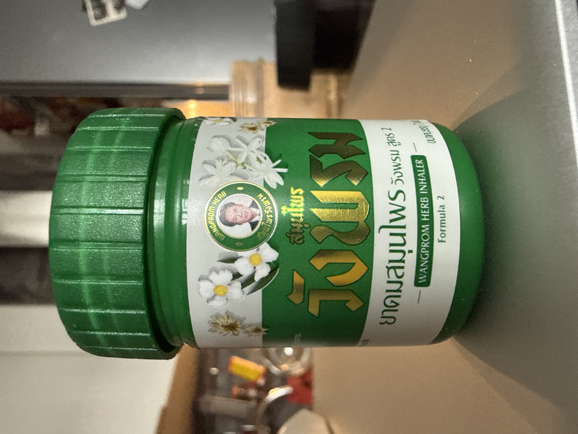An outdoor, eye-level, close-up shot shows a green Wangprom Herb Inhaler, Formula 2, on a metallic surface. The inhaler features white flowers, a portrait of a man, and Thai text in yellow. The background is blurred, showing hints of a kitchen or workspace. No specific location in Domodedovo, Russia, can be determined.