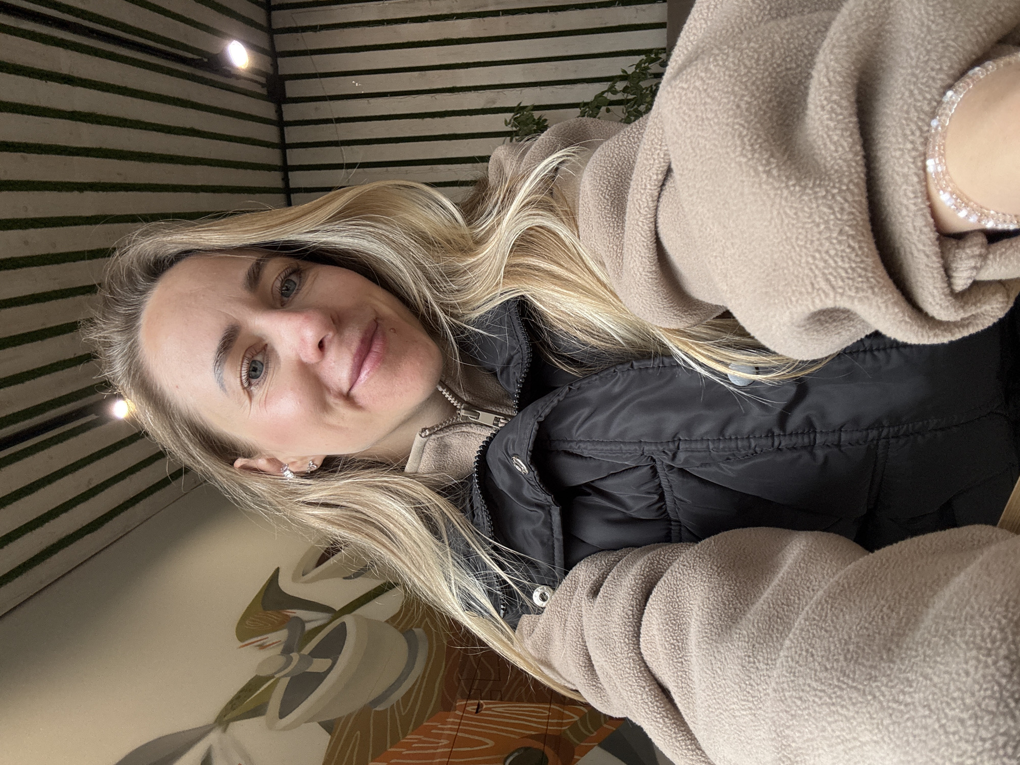 This is a close-up, front-facing selfie of a fair-skinned female individual taken indoors, presumed to be in the city of Chaikovsky, Russia. The woman has long, light blonde hair that falls over her shoulders and displays blue eyes and a subtle smile. Her left ear features multiple earrings, including a stud and a small hoop. She wears a light beige fleece quarter-zip jacket, over which she layers a black puffer vest with visible silver snap buttons. A light pink or clear multi-strand beaded bracelet is present on her right wrist. The background on the left side of the image features a mural or painting on a light-colored wall, depicting stylized elements resembling bowls or cups in shades of brown, beige, and orange. The background on the right, and a portion of the ceiling, consists of vertical green and light-colored stripes. An illuminated overhead spotlight fixture is visible on the right side of the striped ceiling, and a plant with green foliage is partially observable in the far right background.