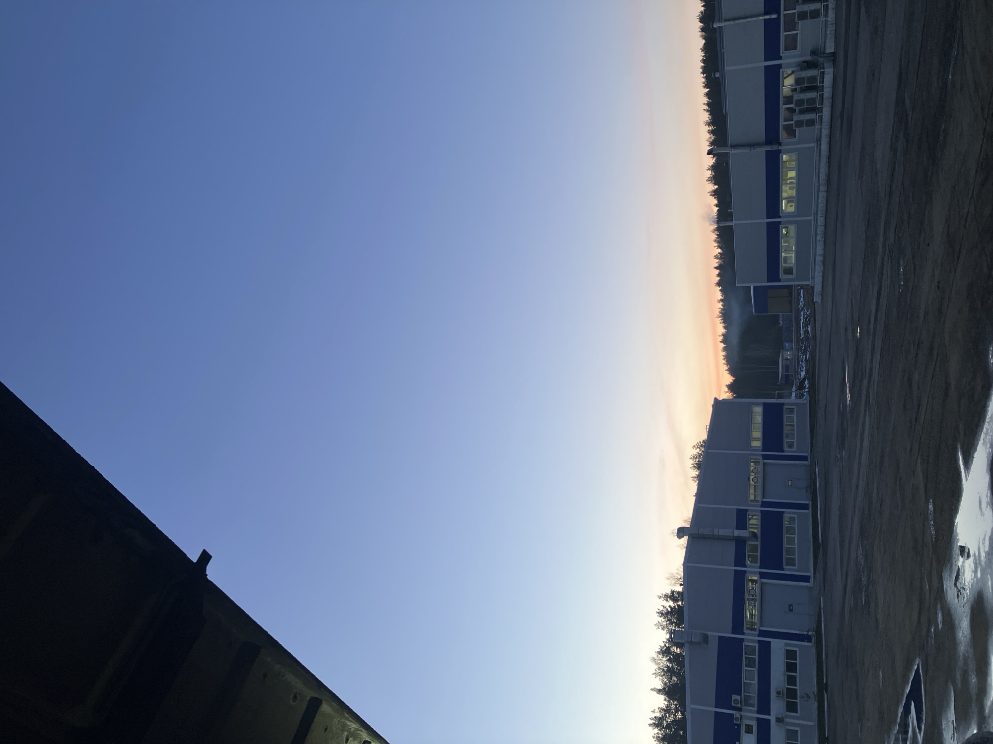 The image depicts an outdoor scene in Noviy, Russia. The sky transitions from blue to a pale orange near the horizon. A row of trees lines the horizon, separating the sky from a row of buildings with light gray walls and blue accents. These buildings have multiple windows, some of which are lit up. The foreground appears to be a dirt or gravel lot, with some areas appearing wet or icy. In the lower-left corner, there is a portion of a dark object, possibly part of a vehicle or building overhang, silhouetted against the sky.
