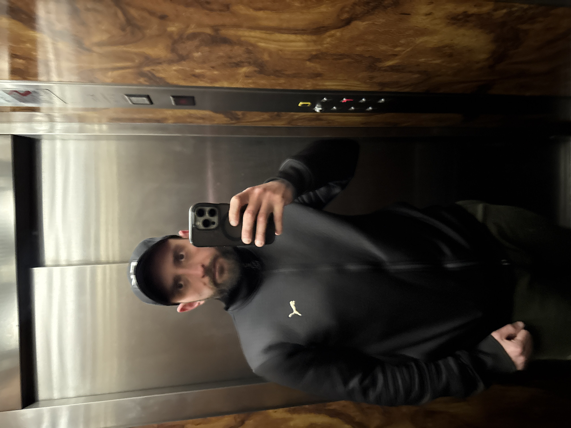 A male individual is observed taking a selfie inside an elevator located in Soria, Spain. The man has a dark beard and mustache, wears a black baseball cap with light lettering, a black full-zip track jacket featuring a light-colored Puma logo on the left chest, and dark pants. He holds a dark-colored smartphone with a triple camera array in his right hand, aiming it towards himself for the photograph. His left hand is clenched into a fist at his side. The elevator interior features a reflective metallic panel on the left (likely the door) and a polished wooden panel with a distinct grain pattern on the right. A metallic control panel column is positioned vertically between these surfaces, displaying several buttons. A red button is illuminated, and a yellow button is also visible. A red indicator, possibly an arrow or overload warning, is faintly visible at the top of the control panel.