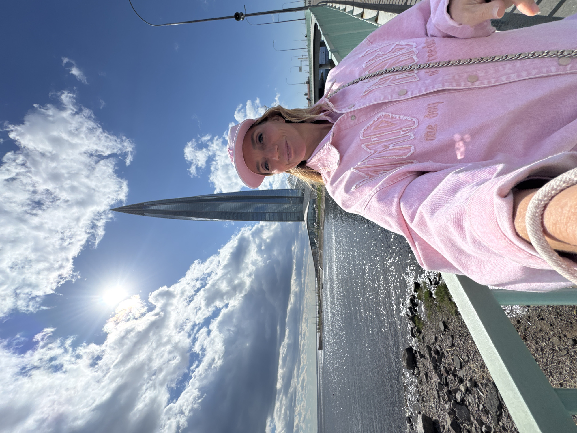 A person is posing for a selfie on a bridge in Saint Petersburg, Russia. The Lakhta Center skyscraper is visible in the background, along with the Gulf of Finland. The sky is blue with scattered clouds, and the sun is shining brightly. The person is wearing a pink jacket and hat. The bridge has a light green railing. The water is calm with sun glinting off the surface. Rocks and vegetation are visible along the shoreline.