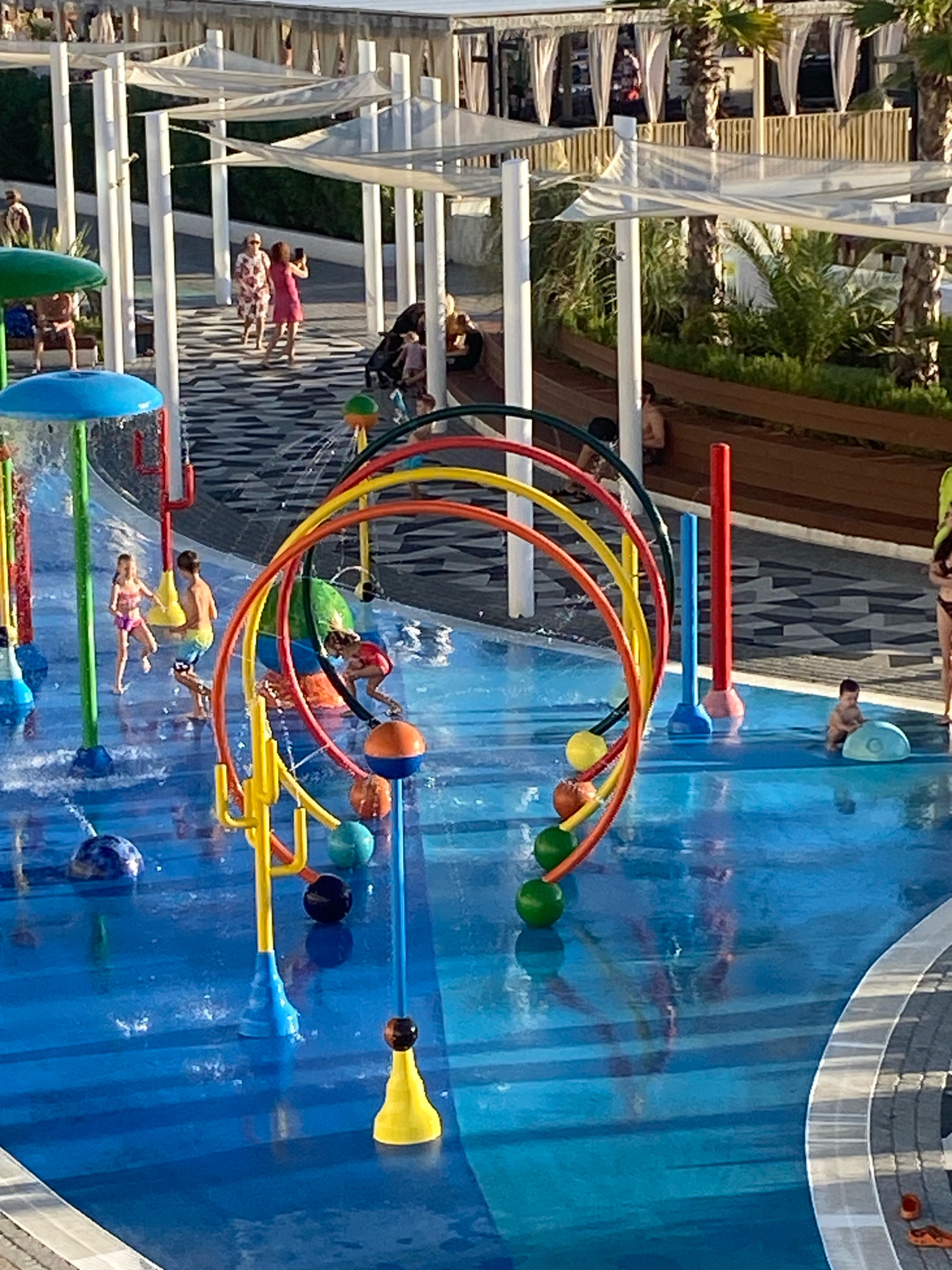 This image depicts a splash pad area, likely at the Sky Park in Sochi, Russia, given the architectural style and tropical vegetation. The splash pad features a bright blue, water-covered surface with various water play structures. In the foreground, a large, colorful, arched structure with red, yellow, and orange rings is prominent, with water spraying from various points. Several spherical, brightly colored water sprayers are scattered across the blue surface.
In the middle ground, two children are visible playing in the water. One child, wearing a pink bikini, is running, and another child, a boy wearing blue shorts, is also in motion. Further back, a toddler is sitting on a blue object in the water, appearing to play.
In the background, a paved walkway with a black and white patterned design runs beneath a covered, open-air structure supported by white pillars and white fabric awnings. Several adults are walking on this pathway, some appearing to take photos. Palm trees and lush green vegetation are visible behind the covered walkway, suggesting a warm climate. The overall scene conveys a recreational and family-friendly atmosphere.