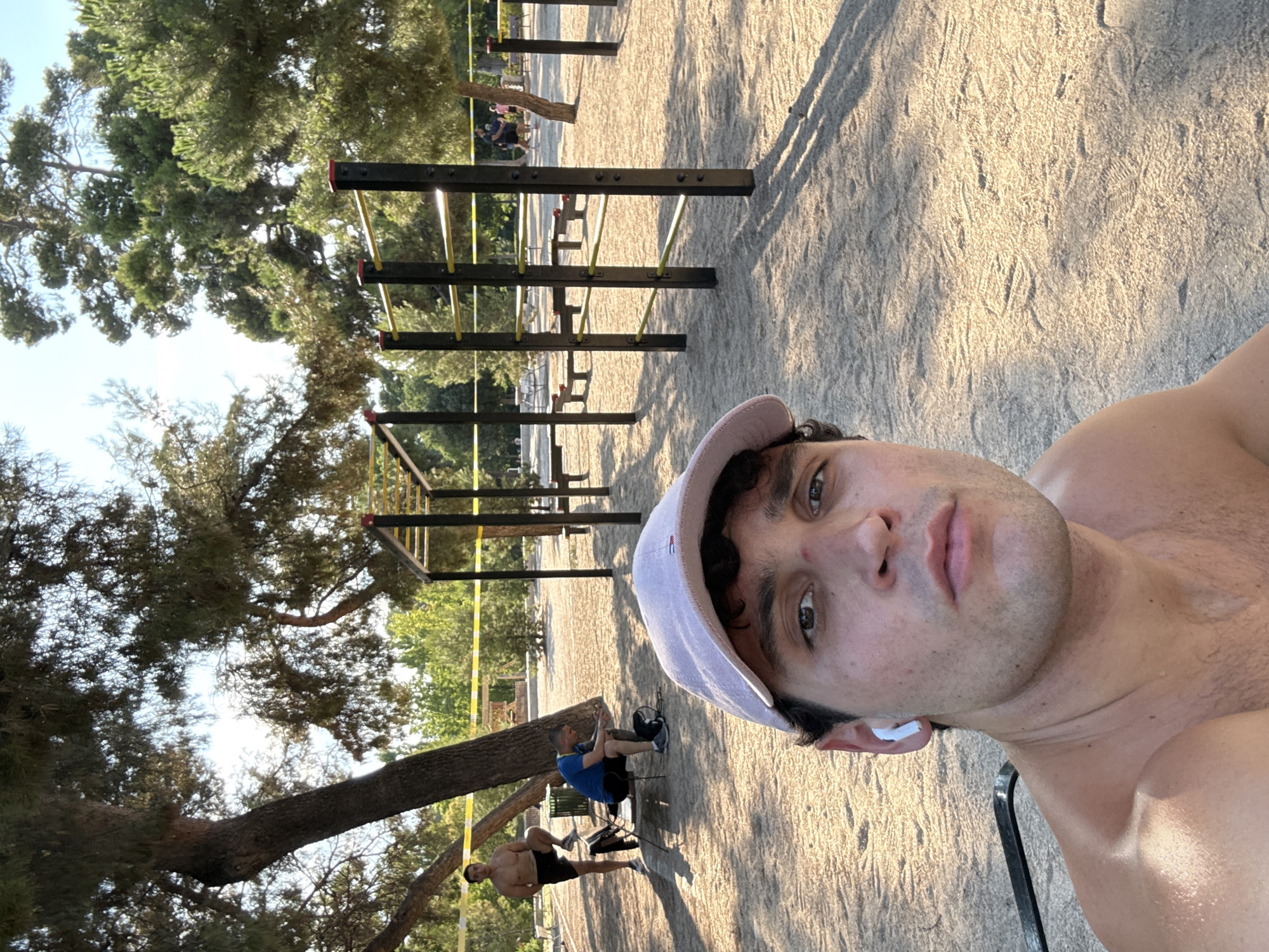 The image depicts an outdoor calisthenics park in Madrid, Spain. In the foreground, a man is taking a selfie, wearing a light-colored baseball cap and white earbuds. He is shirtless and has a fair complexion. Behind him, the park features several structures with horizontal bars, likely for exercises like pull-ups and dips. The ground is covered in sand, and trees with dense foliage are visible in the background, suggesting a sunny day. To the left, two other men are present; one is standing shirtless, wearing shorts, and the other is seated, wearing a blue t-shirt and black shorts. Further in the background, other individuals can be seen, and yellow caution tape is strung between some of the structures. The overall impression is a public space dedicated to outdoor fitness, likely a park within the city of Madrid.