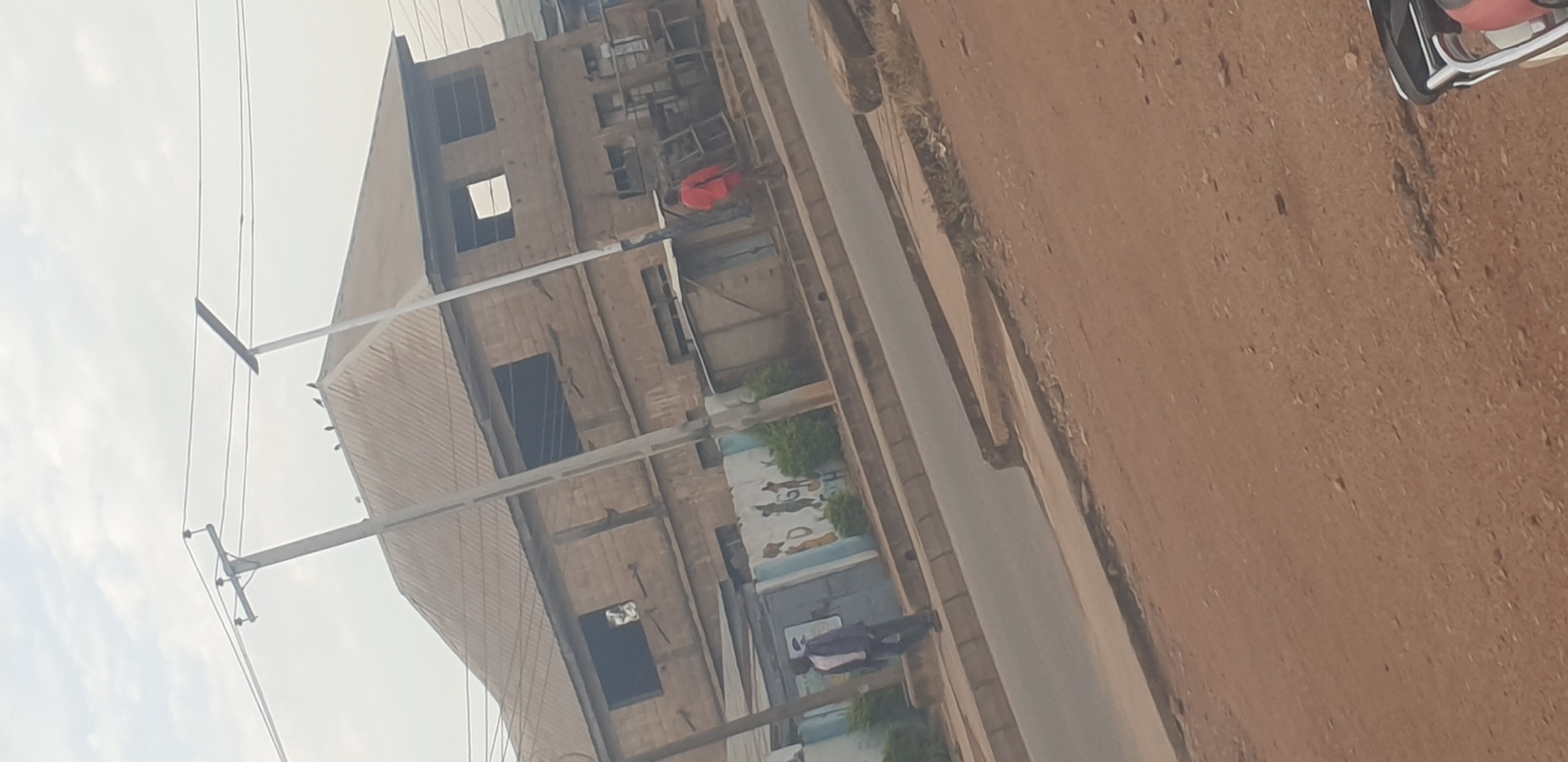 A man in a blue shirt and dark pants walks along a sidewalk next to a two-story building under construction. He is on the left side of the frame. On the right side of the frame, a person in a red shirt walks away from the camera. The building has a corrugated metal roof and brick walls. There are electrical poles and wires visible in front of the building. The ground in the foreground is dirt. The scene appears to be in a city setting, possibly Jos, Nigeria, given the architecture and style of the building.