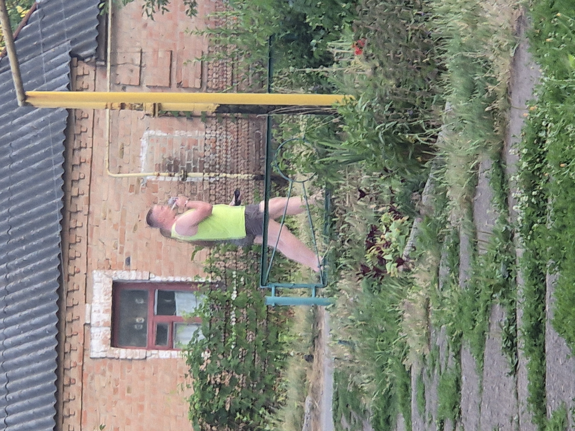 A person is lying on a green metal swing in an outdoor setting. They are wearing a bright yellow tank top and dark shorts. The person appears relaxed, with their head resting on their arm and their eyes closed. The swing is positioned outdoors, near a brick building and surrounded by lush green foliage. A yellow pipe runs horizontally across the scene, attached to the building. The building has a small window with a red frame. The ground is covered with greenery and a path made of stones. The image has a vertical orientation and a soft, slightly blurred quality, suggesting it might have been taken during the daytime with natural light. The overall scene evokes a sense of leisure and tranquility.