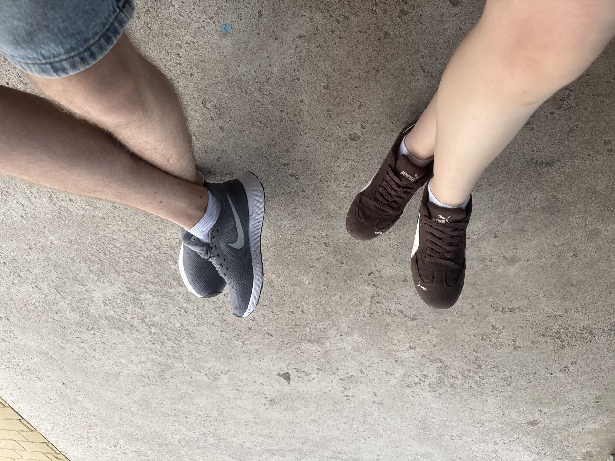 Two pairs of legs are visible, resting on a grey concrete surface. The left legs are wearing brown Puma sneakers with white soles and white socks. The right legs are wearing grey Nike sneakers with white soles and white socks. One person's legs are crossed at the ankles. The concrete is textured and appears to be outdoors, with a hint of a yellow brick-like surface visible in the upper right corner. The lighting suggests it is daytime. No specific location in Shulhynka, Ukraine can be identified from the image alone.