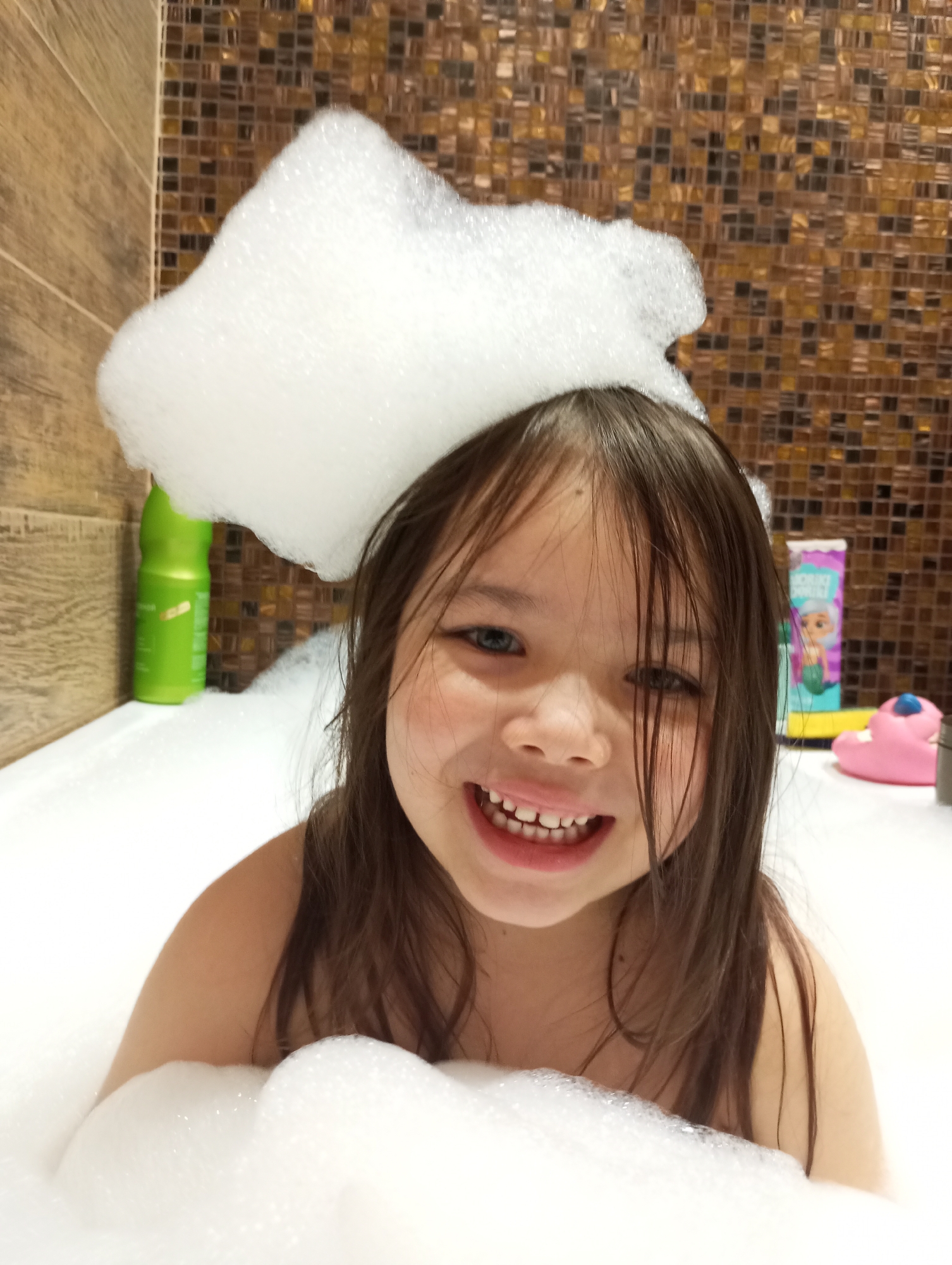 A young girl is in a bathtub full of bubbles. She is smiling and has bubble foam on her head. There are several items on the side of the tub, including a green bottle, a purple and turquoise mermaid-themed container, and a pink toy. The background is a wall with brown and gold mosaic tiles. It is not possible to confirm the location of the image in the city of Pervomaysky, Russia, without additional information.