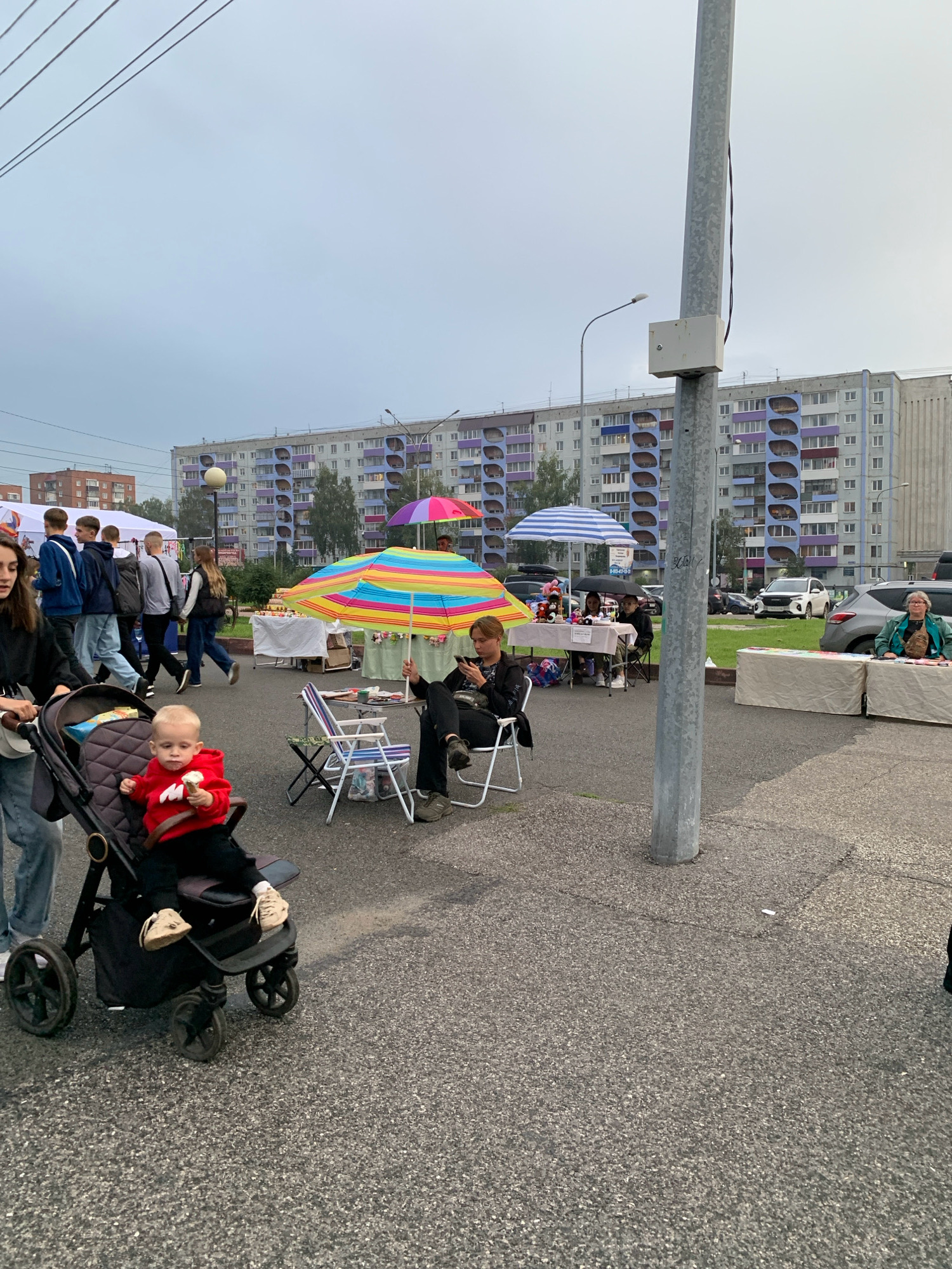 The image depicts an outdoor market scene in Prokopyevsk, Russia, with a large, gray, multi-story apartment building in the background. In the foreground, a child wearing a red sweater is seated in a stroller, holding an ice cream cone. Several people are browsing stalls set up with various goods, some under colorful umbrellas. One person is sitting at a table with an umbrella, looking at their phone, while another individual is seen at a stall on the right. The ground is paved with asphalt, and a tall metal pole with a white electrical box attached stands prominently in the mid-ground. The sky is overcast.