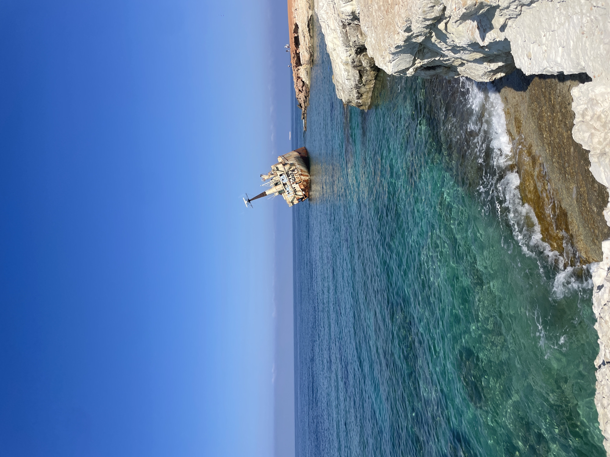 The image depicts the Edro III shipwreck on the coast of Peyia, Cyprus. A rusty, beached cargo ship is tilted dramatically to its port side in the clear, turquoise waters of the Mediterranean Sea. The shipwreck is partially submerged and rusted, with visible damage to its structure. The foreground shows a rocky coastline with white, chalky cliffs and smaller stones leading down to the water. The sea is calm with gentle waves lapping at the shore. In the background, the horizon is clear, meeting a vast expanse of a bright blue sky. A few distant figures can be seen standing on the reddish-brown rocks near the shipwreck, appearing small in the overall scene. The overall environment is a sunny, coastal landscape.