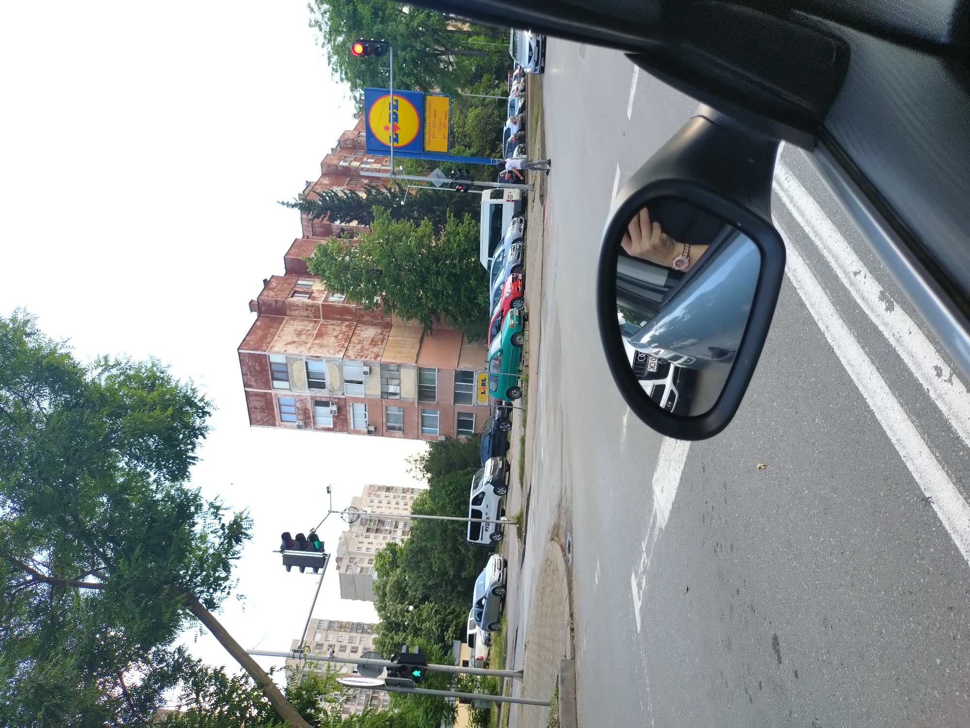 The image, taken from inside a vehicle, captures a bright daytime urban street scene in Stara Zagora, Bulgaria. The perspective looks out the passenger side window and into the right side mirror.
On the right side of the main view, a red traffic light hangs above a prominent blue and yellow circular "LIDL" sign. The sign also displays opening hours in Cyrillic script: "Пон - Съб: 7ч - 21ч / Нед: 8ч - 20ч" (Monday - Saturday: 7am - 9pm / Sunday: 8am - 8pm). Below the sign, a line of cars is parked along the sidewalk, including a white van and several other vehicles. A person in a white shirt and dark pants is walking on the sidewalk near the parked cars. Behind the sign and parked vehicles, a large, multi-story residential building with weathered, reddish-brown facades is visible, interspersed with lush green trees.
To the left, another set of traffic lights shows a green signal, with more residential buildings and extensive greenery in the background. The sky is bright white, indicating clear weather.
In the immediate foreground, the dark frame of the car's interior is visible. The right side mirror reflects a portion of the road with white lane markings, and the driver's hand, adorned with a rose-gold watch, resting on the steering wheel. A partial reflection of a license plate with "O CK" or similar text is also visible in the mirror. The presence of the red traffic light suggests the vehicle is stopped.
