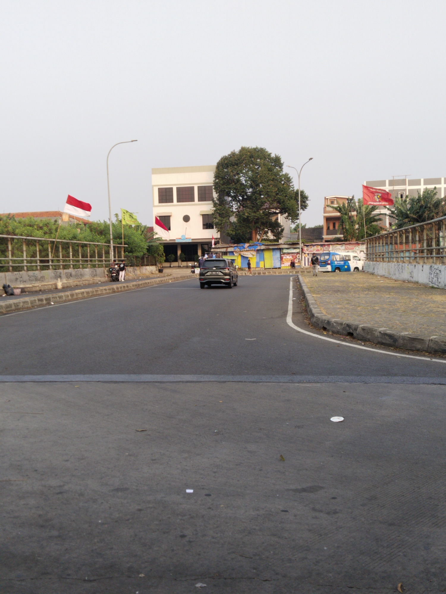 The image depicts a street scene in Depok, Indonesia. The foreground shows a grey asphalt road with white dividing lines. On the right side of the road, there is a raised sidewalk with a patterned brick surface and a concrete curb. Beyond the sidewalk, a metal railing with graffiti marks the edge of an elevated area. In the background, several buildings of varying sizes and architectural styles are visible. Two Indonesian flags, red and white, are prominently displayed, one on the left side of the road and another on the right. There are also other flags, including a red flag with text and a yellow flag with an emblem. A large tree with lush green foliage stands near the center of the image. Several vehicles, including a black SUV and two vans (one blue and white, the other white), are parked or driving on the road and sidewalk area. A few individuals are present, standing or walking near the sidewalk and buildings. The sky is overcast and a pale grey. The environment appears to be an urban setting with a mix of commercial and residential structures.