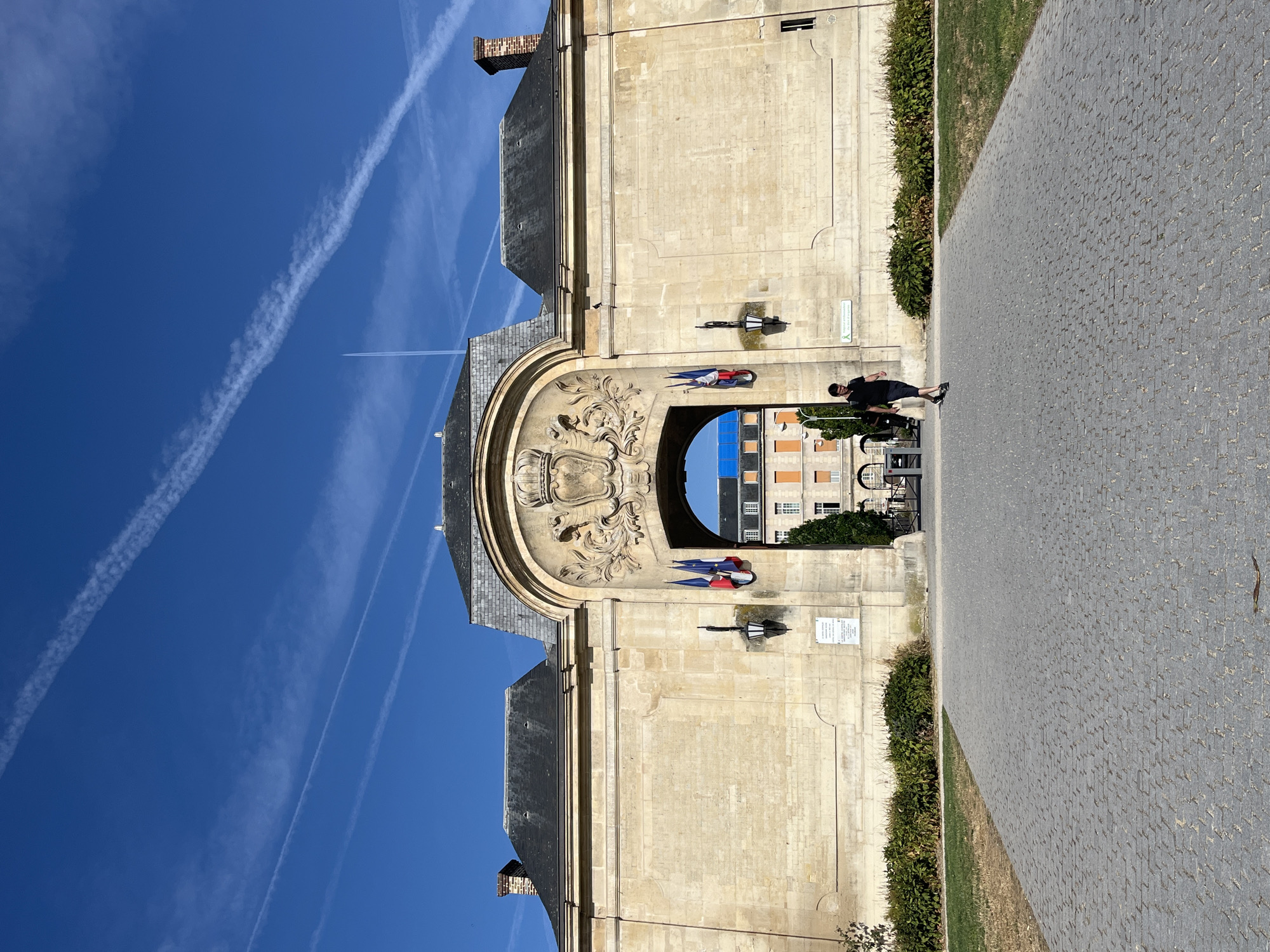 The image depicts a sunlit day in Le Kremlin-Bicêtre, France, featuring the entrance archway of a stone building. The archway is adorned with a crest and two French flags. A lone individual, a person wearing a dark t-shirt and shorts, walks away from the camera on a cobblestone pathway leading through the arch. To the right of the pathway, manicured green bushes line the stone wall of the building, and to the left, a patch of grass is visible. In the background, through the arch, other buildings with orange and white facades can be seen under a bright blue sky with faint white contrails.