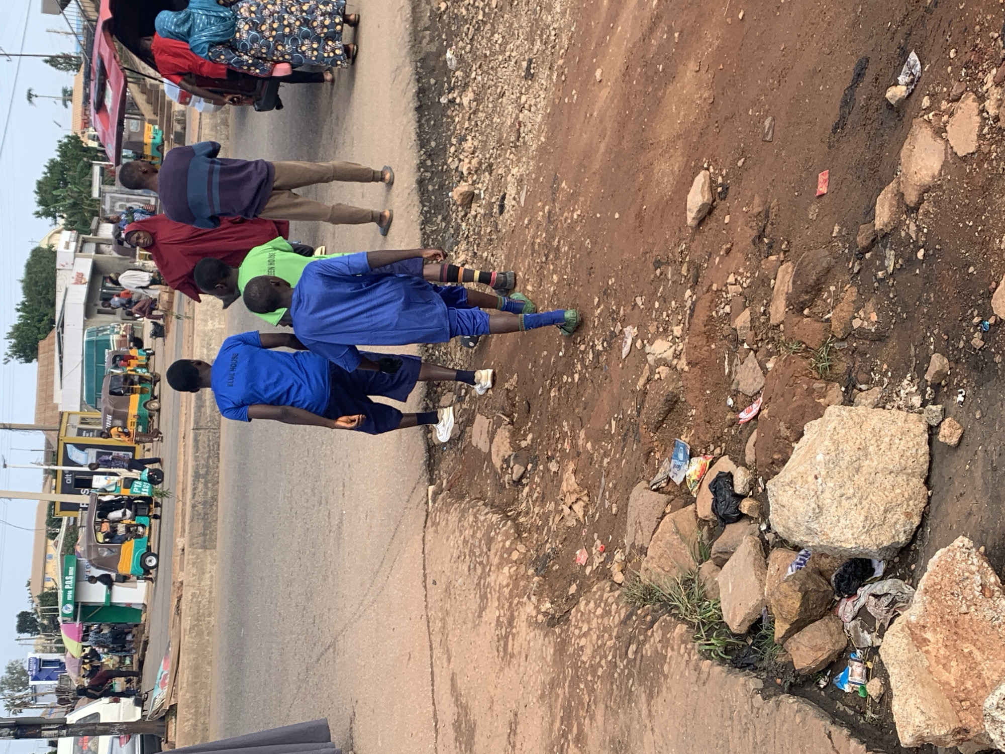 In the city of Gura Top, Nigeria, the image captures a street scene at ground level. In the foreground, the ground is uneven, characterized by a mix of dirt, stones, and debris, with some green vegetation growing between the rocks. A large, light-colored boulder is prominent on the right side of the frame. Further back, a paved road is visible, with several people walking.
On the road, a group of young boys, dressed in athletic wear, are walking away from the camera. One boy in a bright blue t-shirt and dark shorts is in the lead, followed by another in a green t-shirt and dark shorts. Behind them, a boy in a blue t-shirt and blue shorts walks, with another boy in a blue t-shirt and dark shorts to his left. The boys' attire suggests they are likely engaged in some form of outdoor activity or are simply walking in their casual clothes.
Further in the background, several adults are present. A woman in a burgundy abaya stands with her back to the camera, and a man in a striped long-sleeved shirt and brown pants is walking beside her. To the right, a woman in a blue and patterned dress is seen. The street is lined with various structures, including market stalls with colorful umbrellas and what appear to be shops. Several motorized tricycles, commonly known as "keke napeps" in Nigeria, are parked or in motion, indicating a bustling transportation hub or a busy market area. The presence of these tricycles suggests a typical mode of transport in the region. The overall environment depicts a lively street scene in a Nigerian city, with a mix of people, vehicles, and market activities.