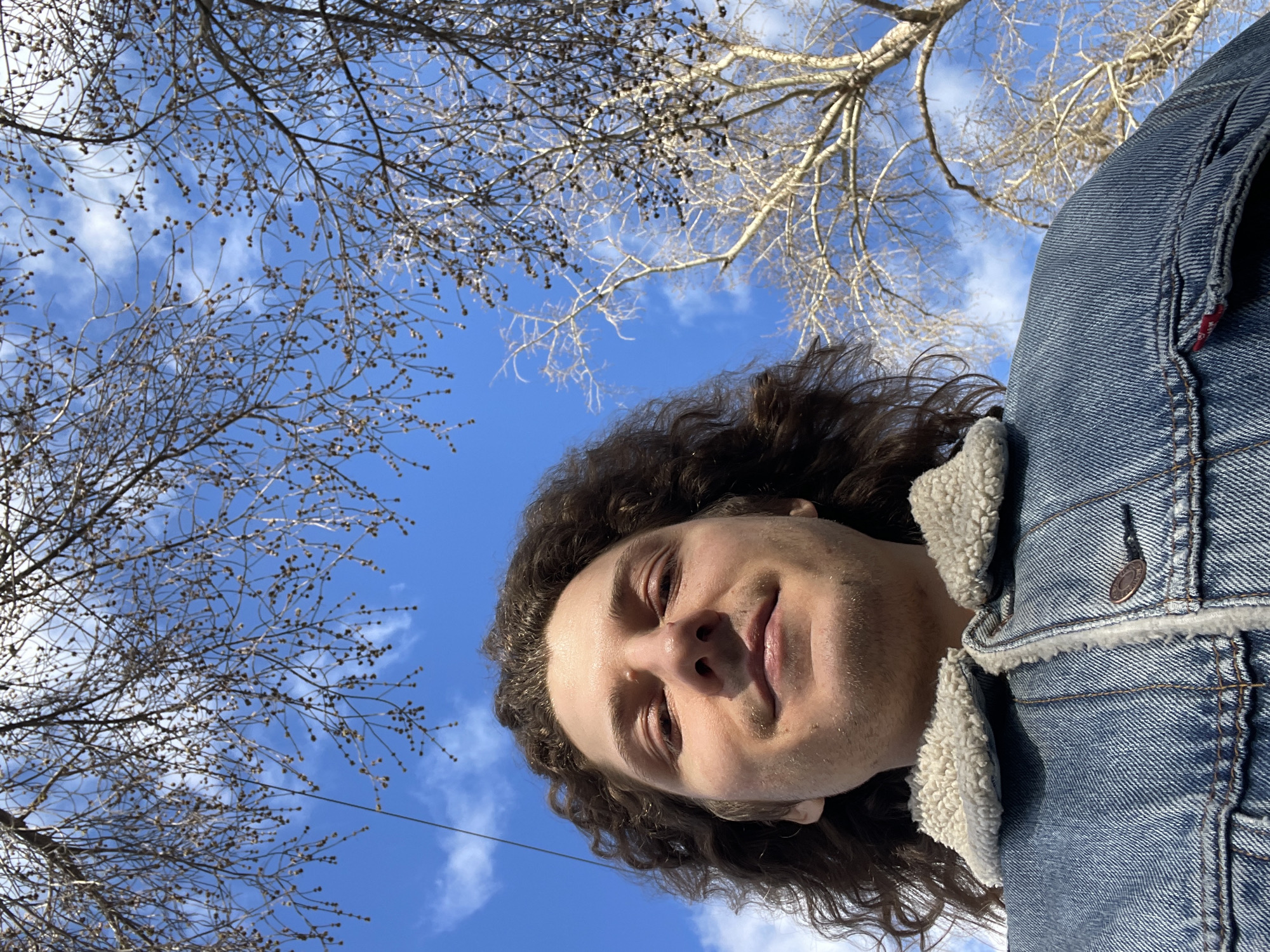 A low-angle shot depicts a fair-skinned individual with long, dark curly hair, wearing a blue denim jacket with a light-colored fleece lining. The person is looking down at the camera. Behind the person is a bright blue sky with scattered white clouds and bare tree branches with small buds on them. The location cannot be determined from the image.