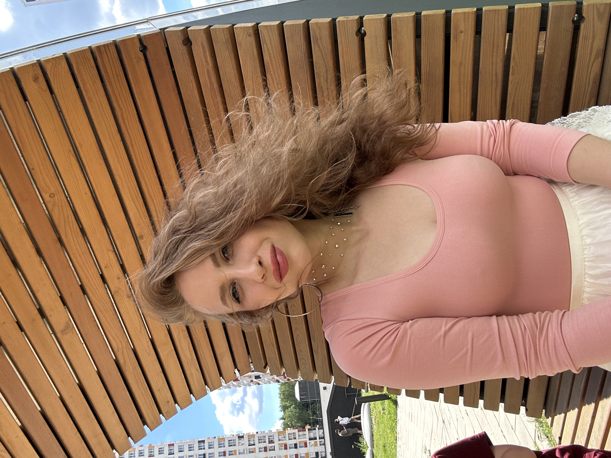 A young woman with long, wavy auburn hair and light skin is captured in a medium shot, facing the camera. She is wearing a pink, long-sleeved top and a white skirt. She has on large hoop earrings and a necklace with rhinestones. The woman is seated on a wooden bench outdoors. Behind her is a wooden architectural structure with horizontal slats, and beyond that, a modern apartment building and trees under a cloudy sky. The location is identified as being in the city of Krasnogorsk, Russia. In the background, two individuals are visible walking on a paved path.