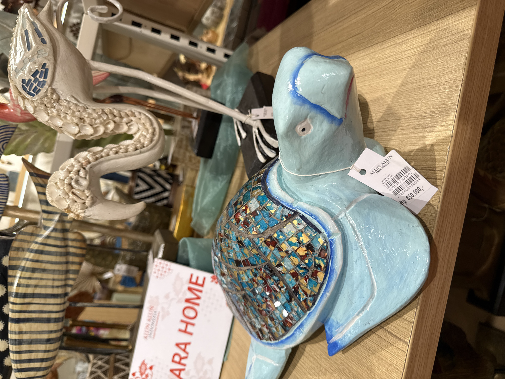 A light blue turtle figurine with a mosaic shell is positioned on a wooden surface. The turtle is facing to the right, with its head slightly lowered. The mosaic shell features a pattern of blue, gold, and red tiles. A white price tag is attached to the turtle with a string. In the background, other decorative items are visible, including a striped fish sculpture and a white flamingo-like figure with shell-like detailing. A red and white sign that reads "ALUN ALUN INDONESIA" and "ARA HOME" is also partially visible. The overall scene appears to be a retail display.