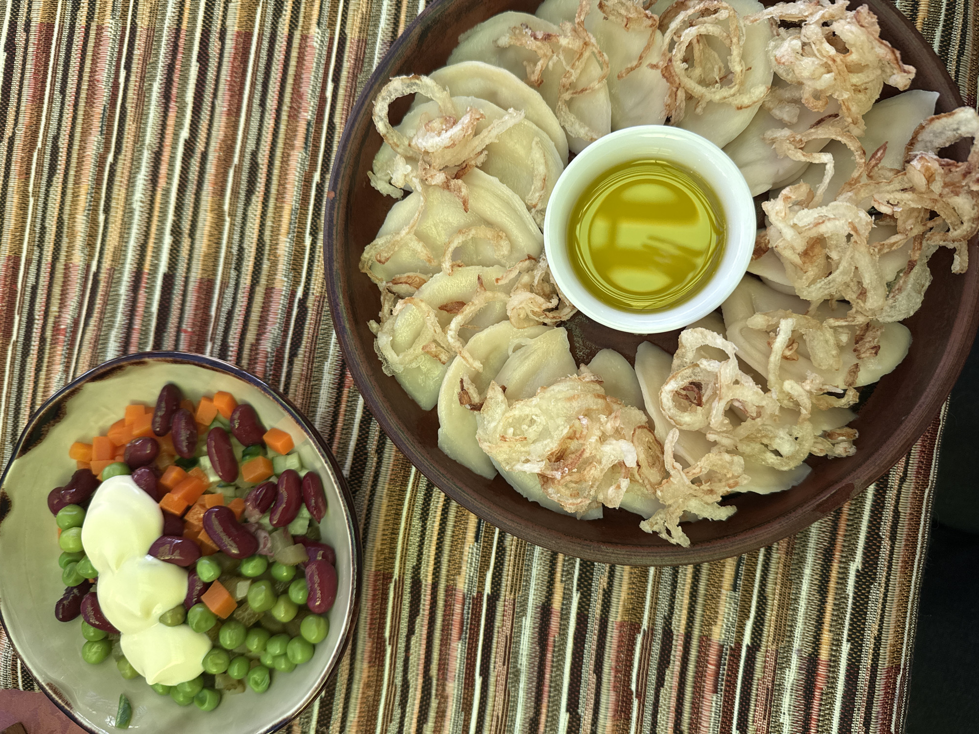 The image shows an overhead shot of two dishes placed on a striped surface. On the left is a small bowl of salad, consisting of peas, kidney beans, diced carrots, and topped with three dollops of white sauce. On the right is a larger, shallow, earthenware bowl containing dumplings. These dumplings are garnished with fried onion rings and accompanied by a small white bowl filled with a yellow liquid, likely oil or sauce. The striped surface appears to be a woven mat or fabric. The location is likely Kladovitsy, Russia, given the prompt's instructions.