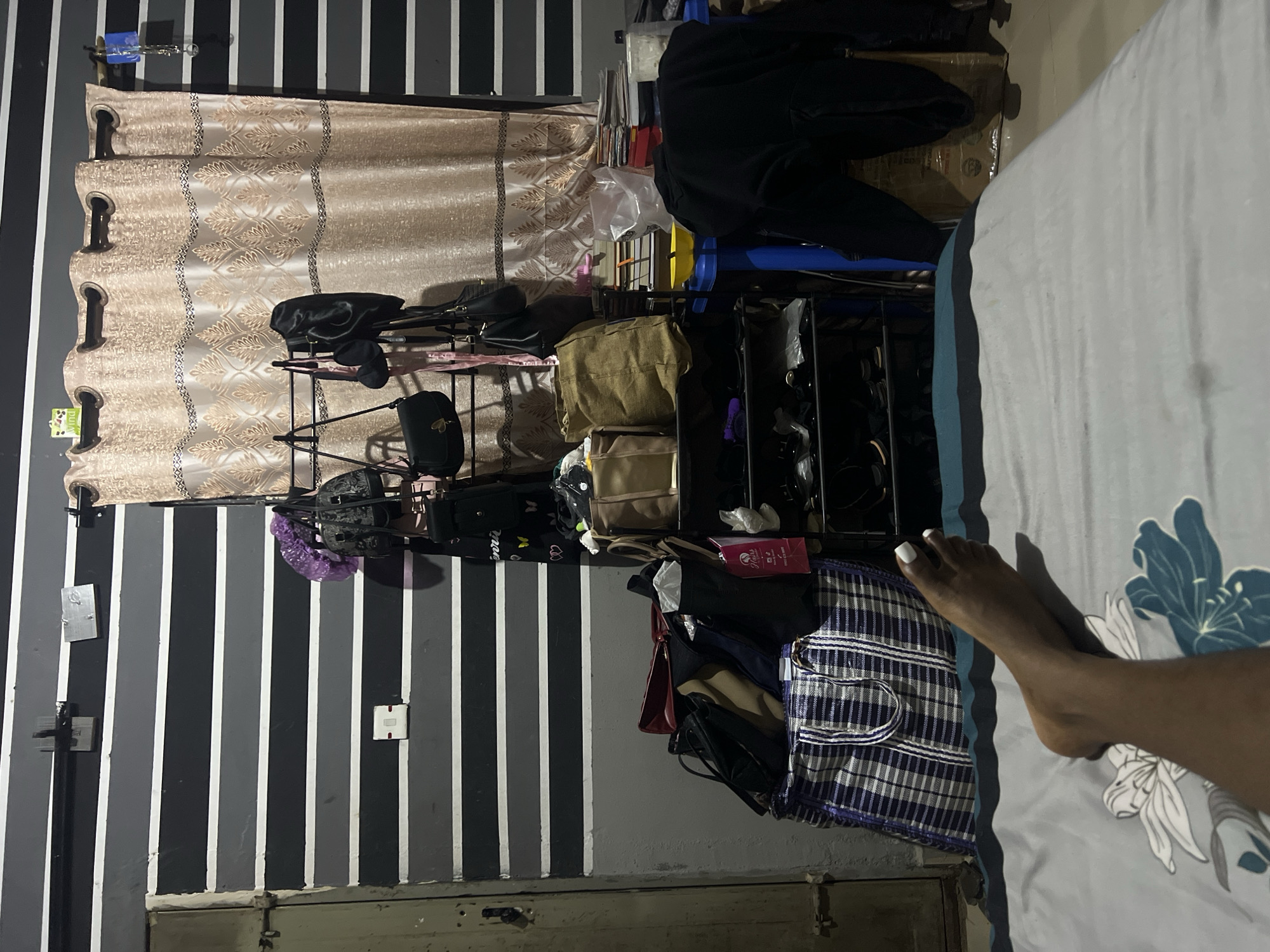 The image shows a bedroom in Jos, Nigeria, at night. The room has striped walls of black and gray, and a cream-colored curtain with a patterned design. In the foreground, a person's bare foot with white-painted toenails is visible on a bed with a floral patterned duvet. The background is cluttered with various items. A shoe rack filled with shoes and bags is visible, along with several handbags hanging on a rack. A large checkered bag is also present. Books, papers, and a blue stool are also visible in the scene. The overall impression is one of a lived-in space.
