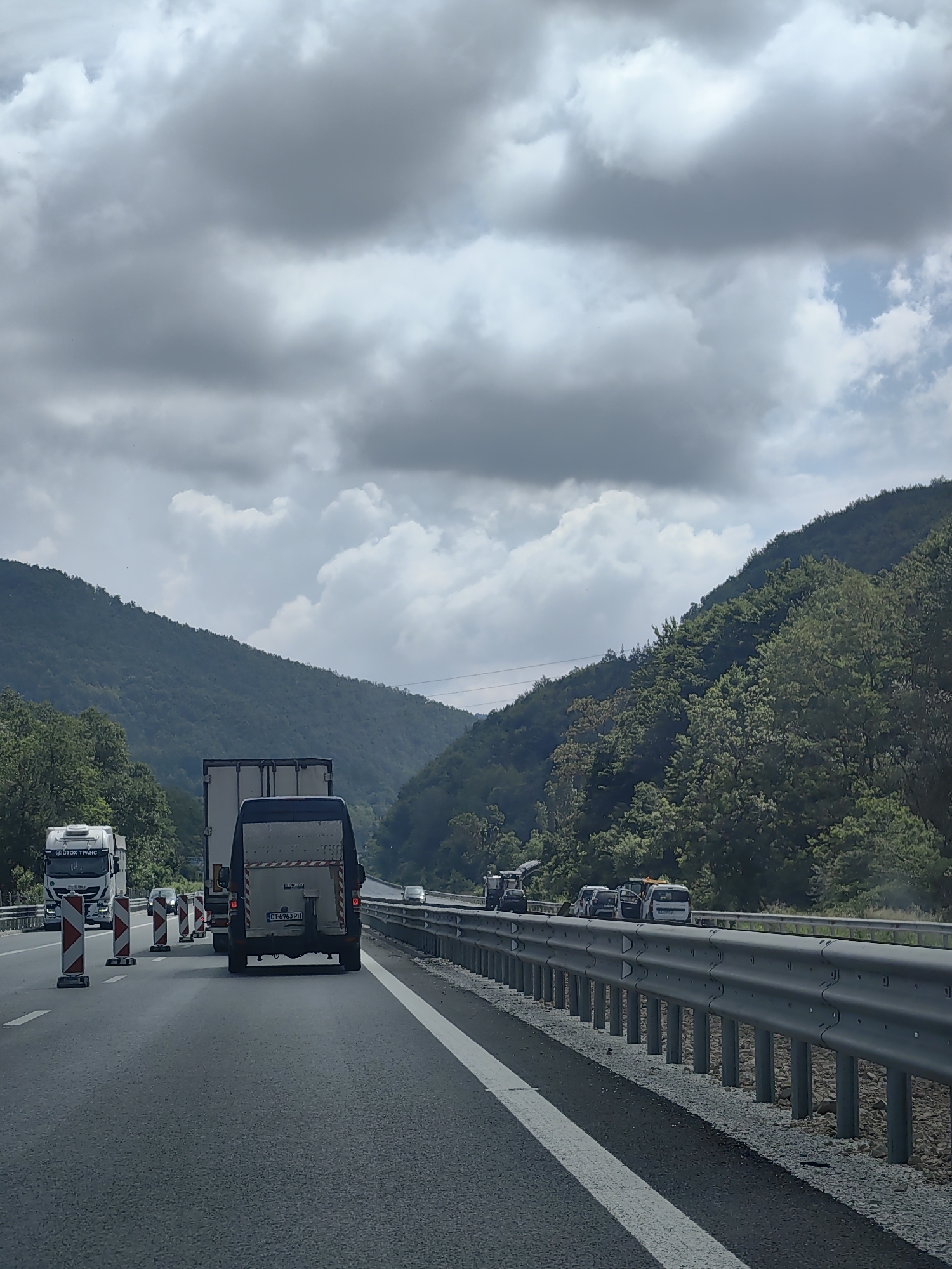 The image captures a view from a vehicle traveling on a highway, likely the A1 highway in Bulgaria, near Churek. The scene depicts a traffic jam with several vehicles, including trucks and vans, stopped on the road. The highway is bordered by a metal guardrail and then a steep, tree-covered hillside on the right. On the left, there are more trees and a less steep, wooded hill. The sky is filled with heavy, gray clouds, suggesting overcast weather. Several construction cones are visible on the road, indicating potential road work. The overall impression is one of congestion and a slightly gloomy atmosphere due to the weather conditions.
