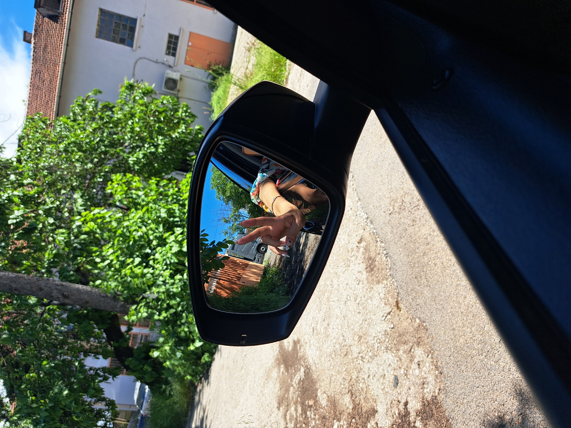 The image shows a view from inside a car, focusing on the driver's side mirror. The mirror reflects a hand giving a peace sign. Behind the hand, the reflection shows a street scene with a red-tiled roof of a building, greenery, and a light-colored building in the background. Outside the car, the road surface is visible. The image was taken during daylight.
There is insufficient information to determine if the image was captured in the city of Sofia, Bulgaria.
