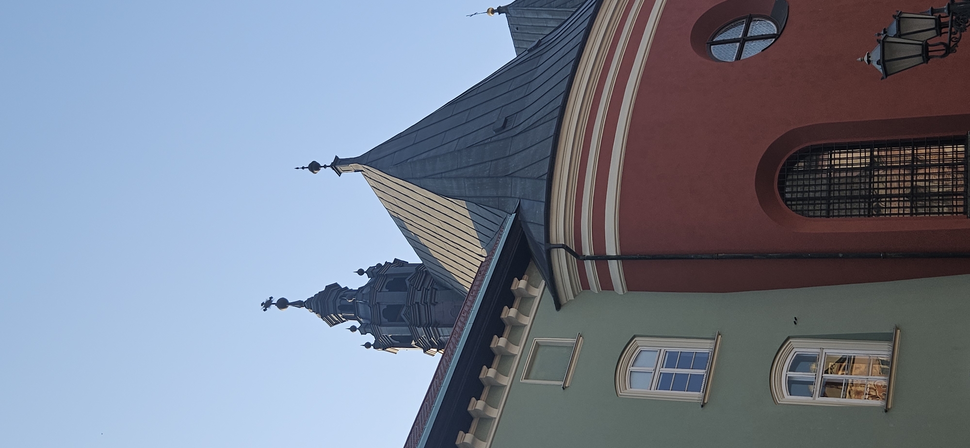 This image shows the facade of a historic building in Krakow, Poland, specifically the side of St. Mary's Basilica. The building features a red section with a large arched window covered by a metal grate and a smaller circular window above it. To the left, a green section of the building has two windows. The roofs are dark and sloped, with a prominent spire topped by a cross. The sky is clear and blue. The architectural style suggests a Gothic or Renaissance influence, typical of Krakow's Old Town. The image is taken from a low angle, looking up at the building.