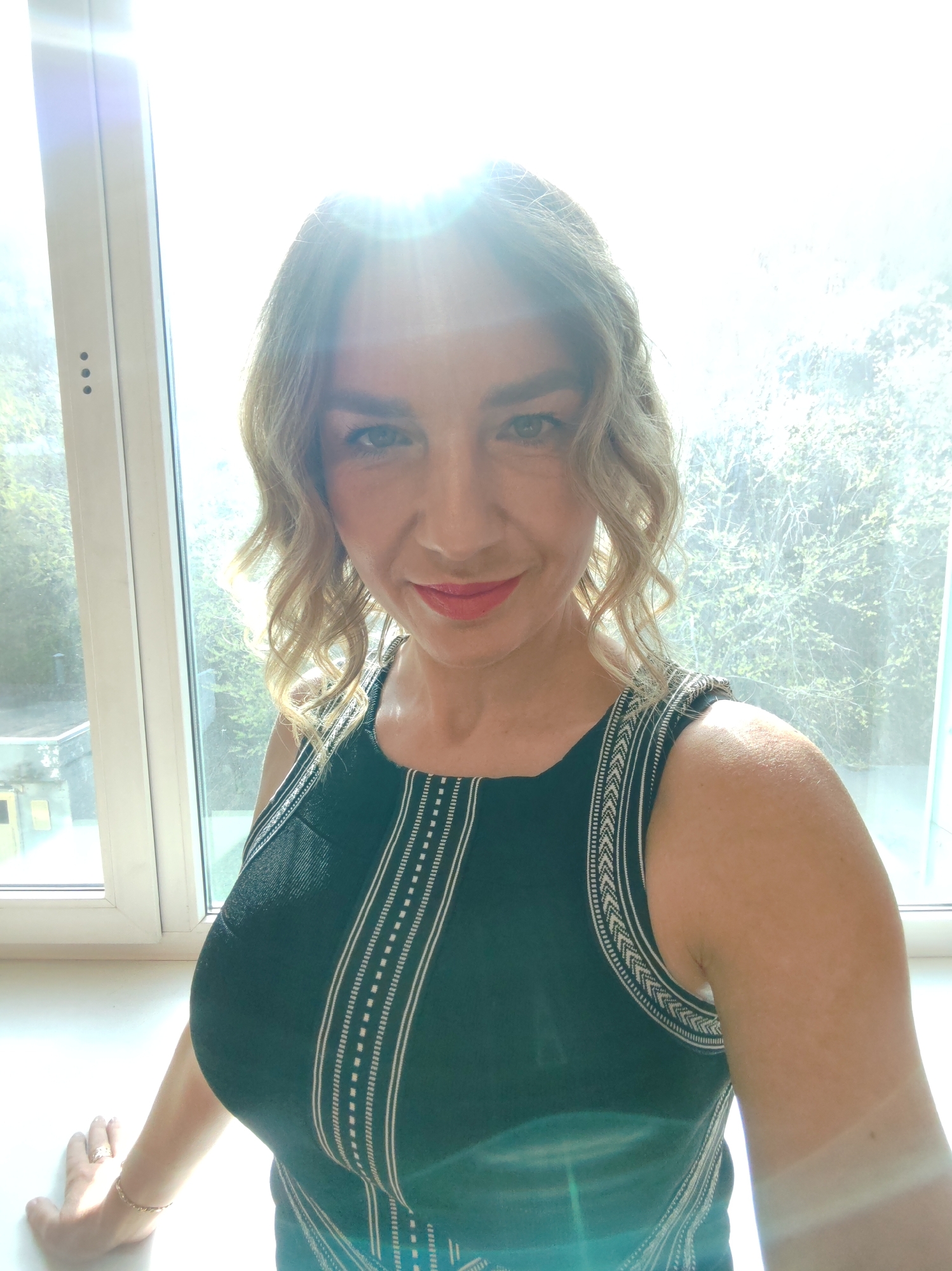 A woman stands indoors, positioned in front of a window. She is wearing a sleeveless black top with white patterned detailing. Her blonde hair is styled in curls. Light streams through the window, creating a bright, almost overexposed environment. The location appears to be an interior space with a window overlooking an area with trees. The city and specific location are unknown.