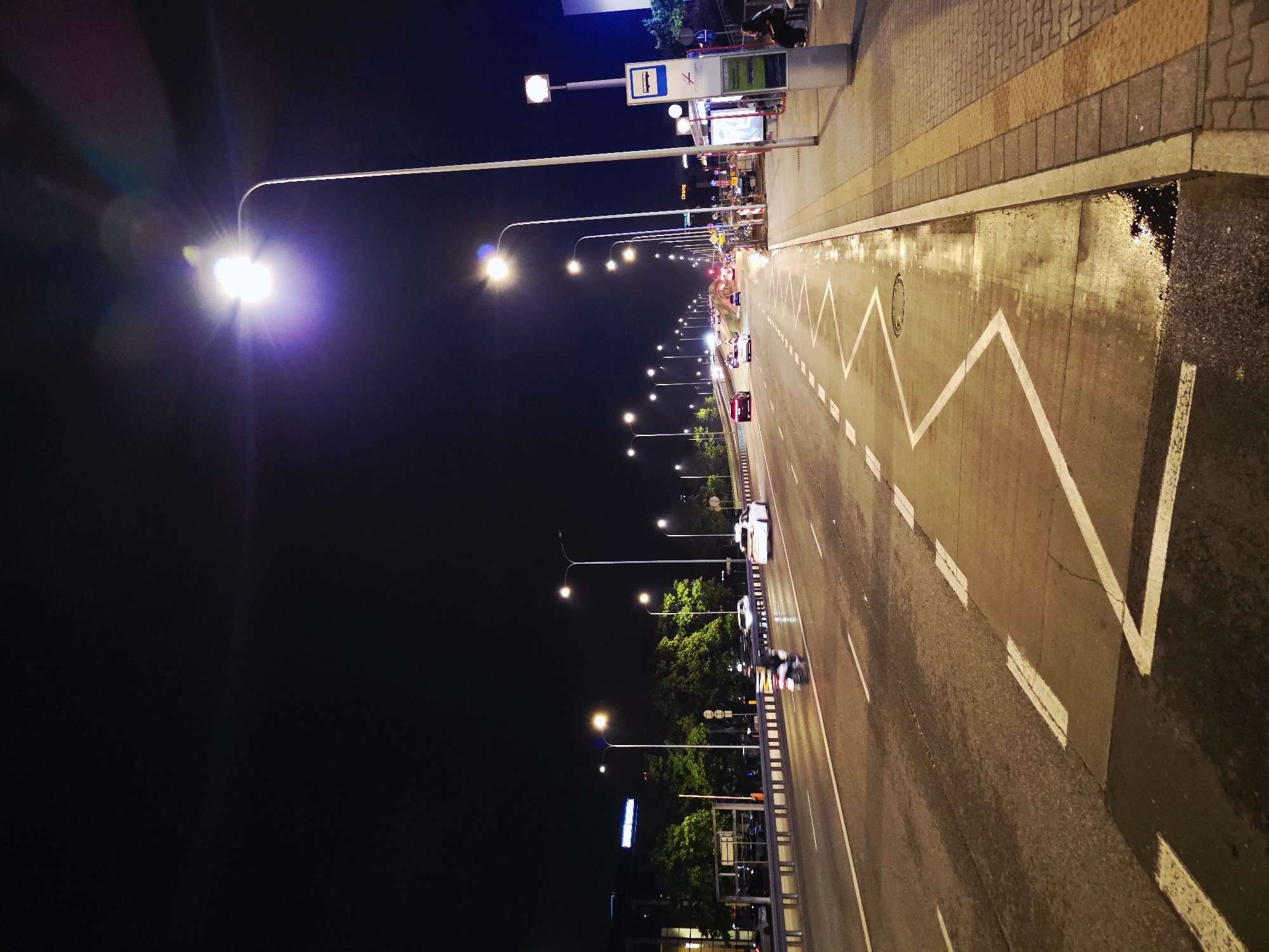 The image displays a night-time scene of an urban road stretching into the distance, illuminated by numerous streetlights. The perspective is slightly elevated, looking down the length of the road.
On the right side of the frame, there's a paved sidewalk with distinct light and dark sections. A bus stop is visible, marked by a sign featuring a blue bus icon and a red no-stopping symbol. A dark, obscured figure can be discerned near a bus shelter behind the sign. The sidewalk edge closest to the road shows a small puddle of water, reflecting the bright artificial lights.
The multi-lane road surface appears wet and reflective in several areas, suggesting recent rainfall. White road markings are prominent, including dashed lane dividers, solid lines, and distinct white chevron or zigzag patterns, typically indicating a no-stopping zone or a specific lane designation. Several vehicles, including cars and possibly a motorcycle, are present on the road, their headlights and taillights creating visible light sources.
Numerous streetlights line both sides of the road, emitting bright, somewhat purple-tinged light that creates noticeable lens flares. On the left side of the road, behind a metal guardrail, lush green trees are visible, illuminated by the streetlights. Further in the background, more artificial lights from distant buildings and other sources contribute to the urban glow against the dark night sky. Some signs near the bus stop have partially readable text, appearing to include characters like 'CYRBO' or similar. The overall scene depicts a functional, active city thoroughfare during the night, likely after rain.