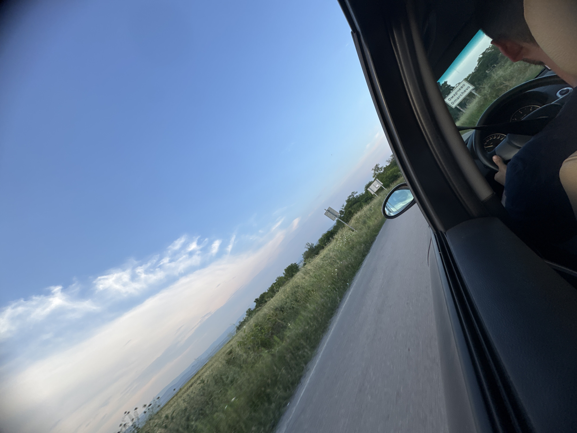 The image depicts a tilted, eye-level view from inside a moving vehicle on a road near Omarchevo, Bulgaria. The driver's arm and part of their head are visible on the lower right, with hands gripping the steering wheel. The car's black dashboard and the top edge of the open window frame the lower and right sides of the image. Through the window, an asphalt road with a faint white line on its edge recedes into the distance, bordered by a wide expanse of green grass and scattered low vegetation. Further back, a line of trees and distant rolling hills are visible. The sky occupies the upper portion, exhibiting a clear blue with streaks of white, wispy clouds stretching across the frame. A white rectangular road sign, bearing the text "Омарчево Omarchevo" in both Cyrillic and Latin scripts, is clearly visible on the right side of the road, slightly ahead of the vehicle. The driver's side mirror is also visible, reflecting a distorted portion of the sky and the road. The lighting suggests daytime, possibly late afternoon, casting an even illumination over the scene.