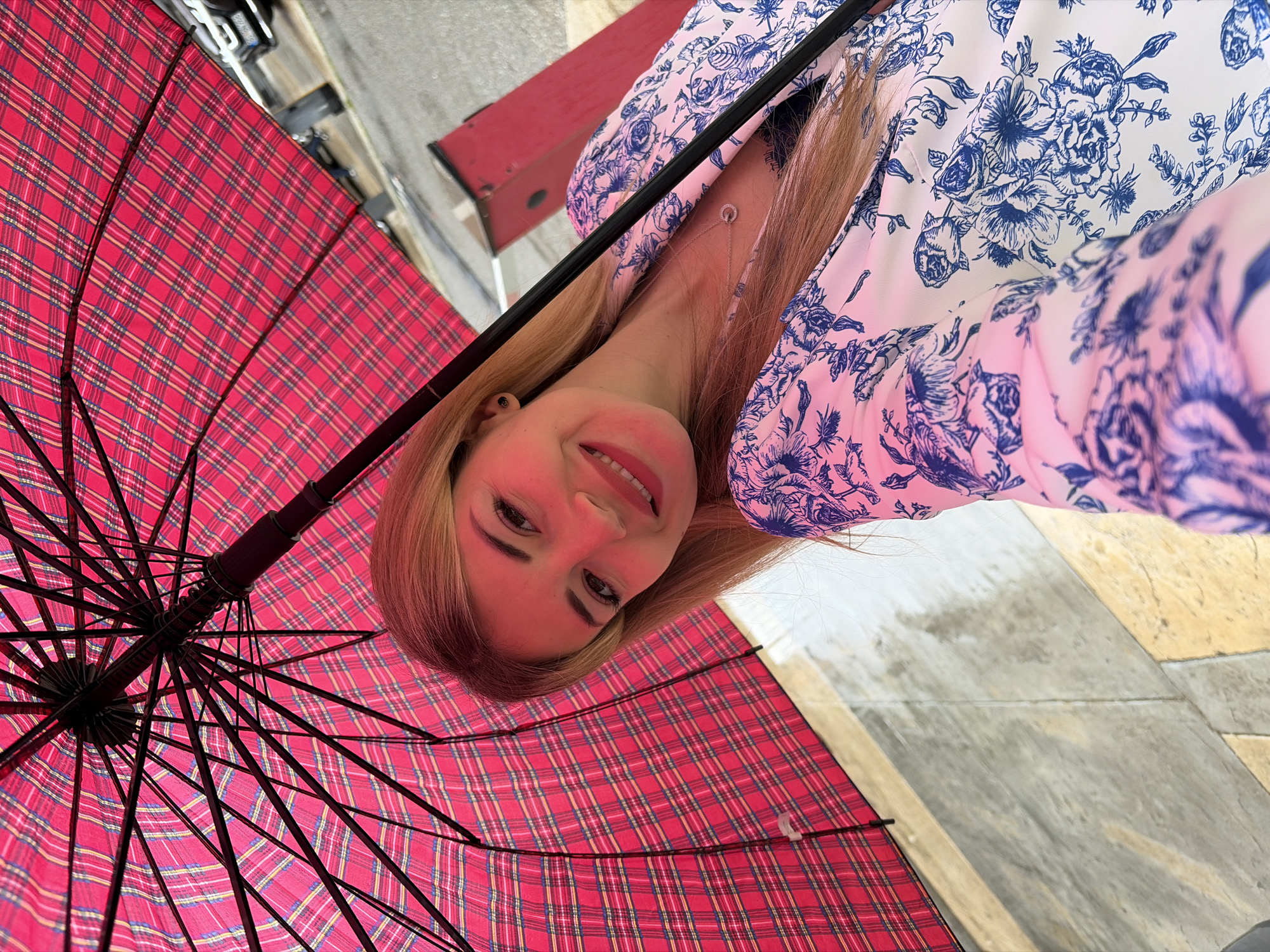 An eye-level selfie shows a smiling young woman holding an open, red plaid umbrella, shot from below/upward. The image is rotated ninety degrees clockwise, so the woman's head is on the left and her body extends towards the right, with the umbrella occupying the left half of the frame.
The woman has long, light brown hair that falls around her face and over her shoulders. She is wearing a white garment with a prominent blue floral pattern. Around her neck is a delicate silver chain with a small, circular pendant. Her face is turned towards the camera, illuminated with a warm, slightly reddish glow likely cast by the umbrella. She has dark eyebrows and eyelashes, and a wide, cheerful smile revealing white teeth.
The umbrella, open and spread above, is red with a distinct plaid pattern featuring intersecting lines of blue, yellow, and red. Its black metal ribs and central pole are visible, indicating it's open. The umbrella covers much of the left side of the image.
To the right of the woman and below the edge of the umbrella, the ground is visible, appearing to be paved with rectangular sections of light tan and grey stone or concrete. In the blurred background beyond the woman's head, there are hints of a street or outdoor environment, possibly with buildings or vehicles. The lighting is bright, suggesting daytime. The woman's expression is joyful and engaging, implying a pleasant moment captured outdoors in Budva, Montenegro. No visible text is present.