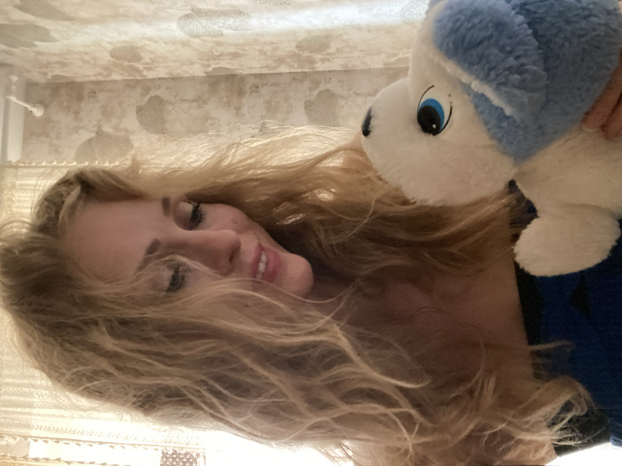 A woman with blonde, wavy hair is holding a white and blue stuffed animal in her arms. She is looking down and smiling. The background appears to be a room with patterned wallpaper and a sheer curtain. The scene is likely indoors, possibly in a residential setting.