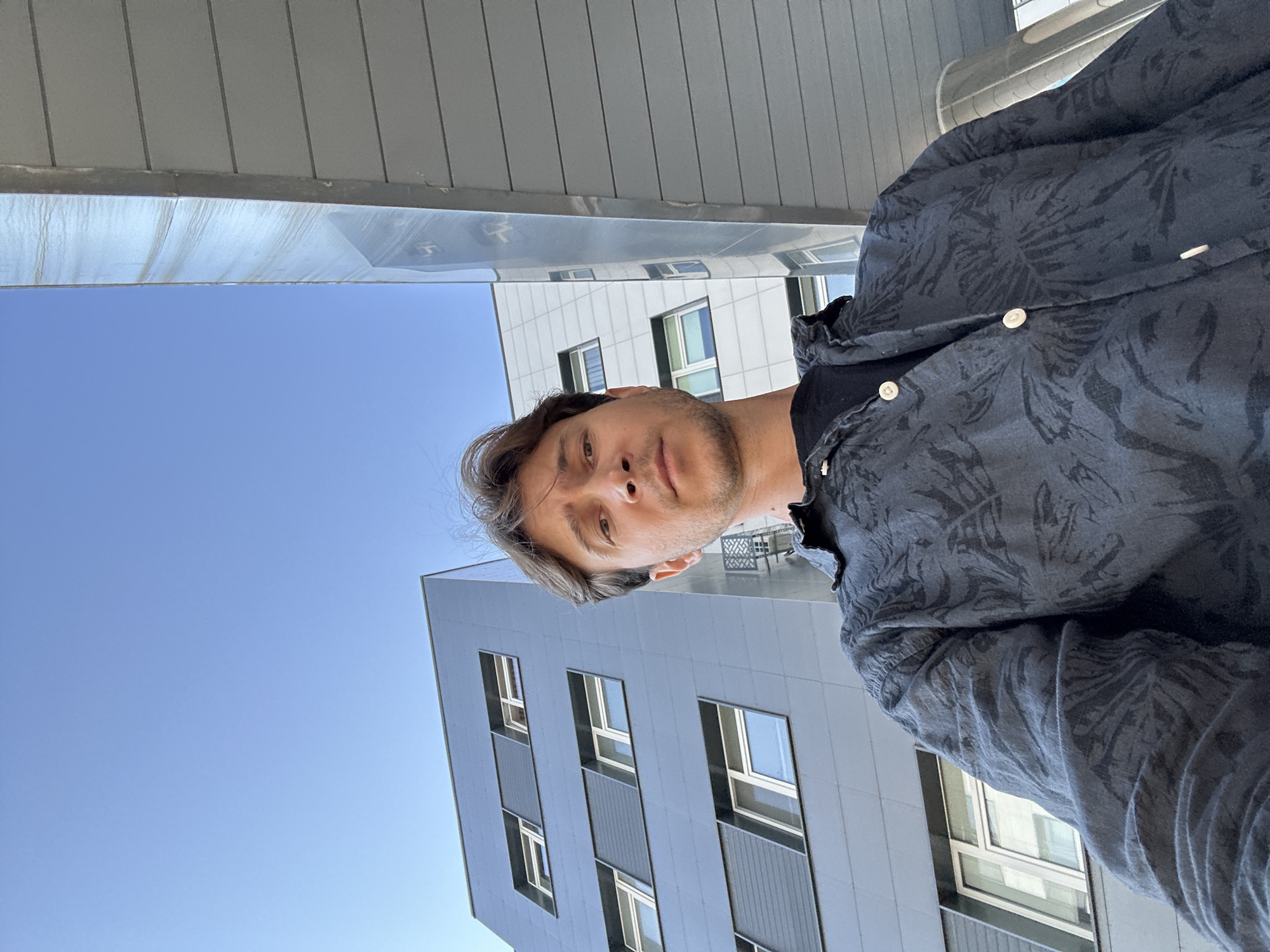 A selfie taken from a low angle shows a man with light brown hair and a neutral expression standing outside. He is wearing a dark patterned shirt over a black t-shirt. Behind him, the facade of a modern building with multiple floors and windows is visible, along with a clear blue sky. The architecture suggests an urban environment, possibly in Kyiv, Ukraine, given the context.