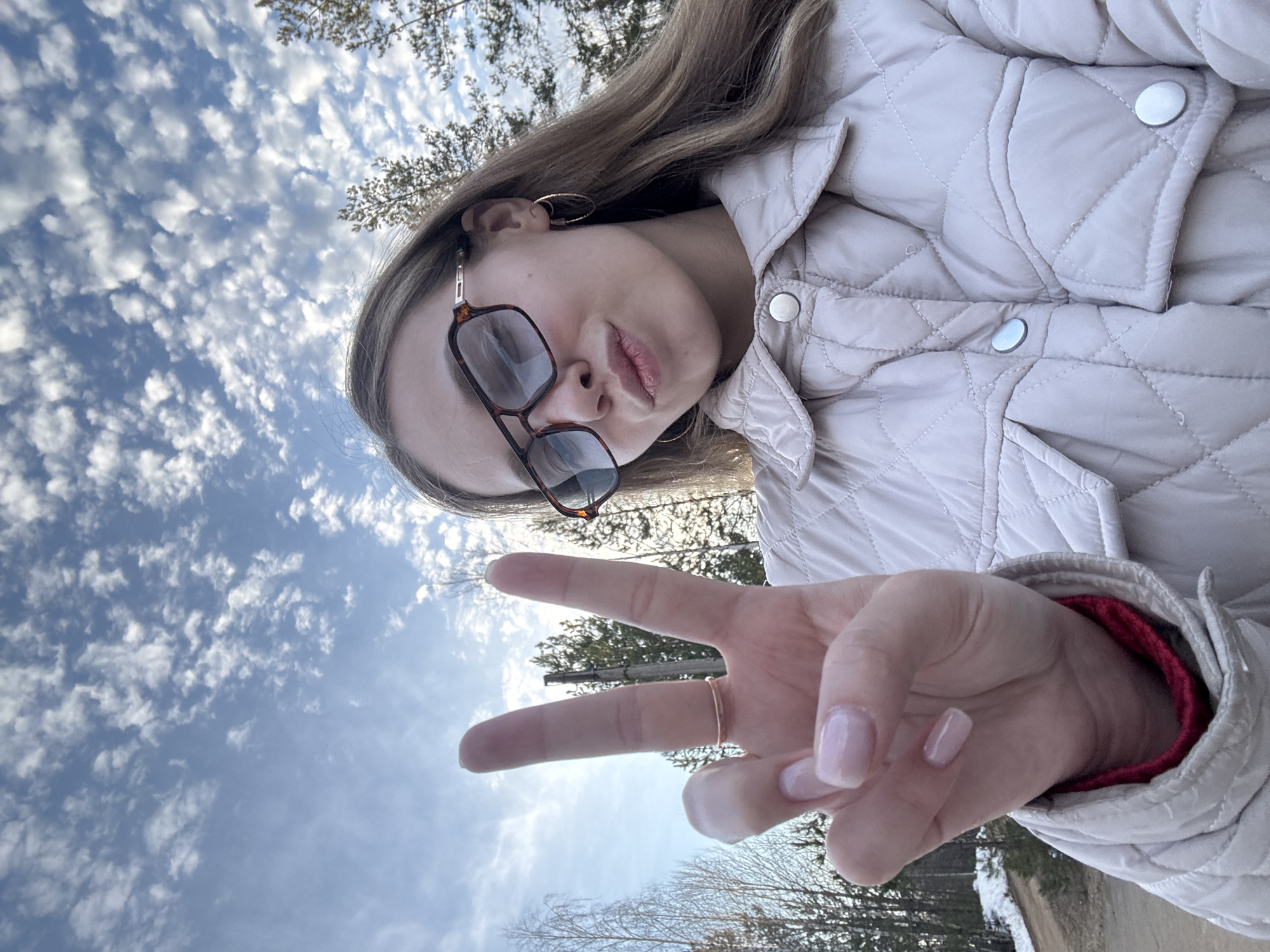 A young woman is taking a selfie outdoors. She wears a quilted jacket, eyeglasses, and hoop earrings. She is making a peace sign with her fingers. The background includes a blue sky with clouds and trees. The location cannot be determined to be in Chaykovsky, Russia without further information.