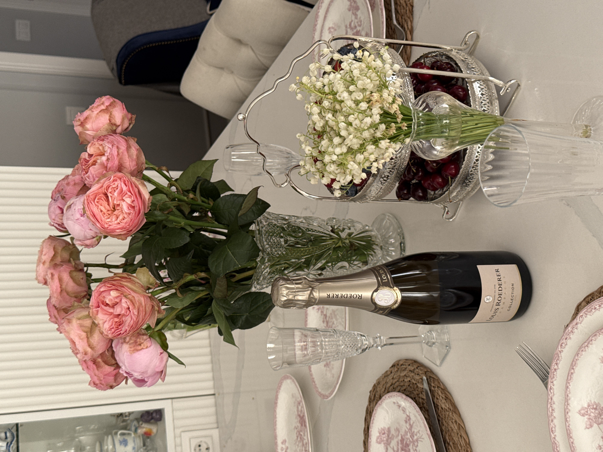 The image captures a dining table set for a special occasion. A bouquet of pink roses is arranged in a vase towards the left side of the table. Adjacent to the roses is a decorative silver stand containing a floral arrangement of lily of the valley and a selection of red cherries and blueberries. A bottle of Louis Roederer champagne rests on the table near a crystal champagne flute. The place setting includes floral-patterned plates with natural fiber placemats and silverware. The setting suggests a celebration or an elegant meal, possibly within a private residence or a restaurant. The specific location in Moscow, Russia is not determinable from the provided image. There are no people visible in the image.